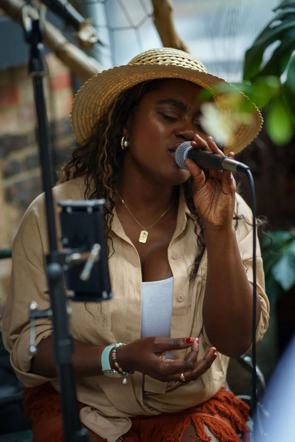 A woman wearing a straw hat, earrings, and necklaces, singing into a microphone with her eyes closed.