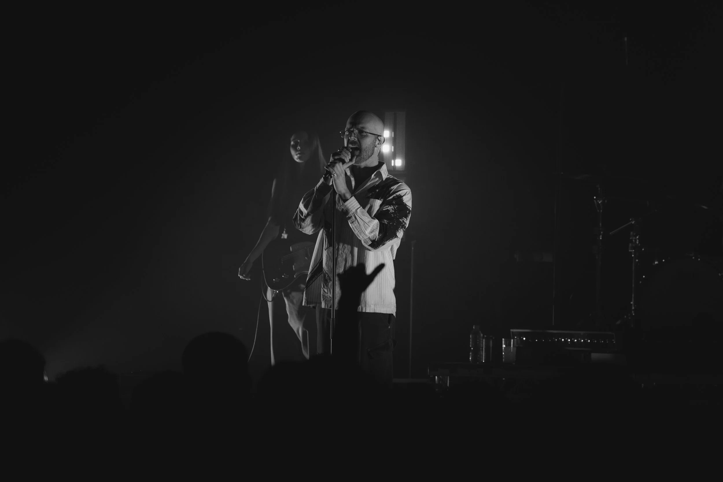 A black and white photo of a man singing into a microphone on stage, with a female guitarist in the background and stage lights behind them, and a silhouette of a hand in the foreground.