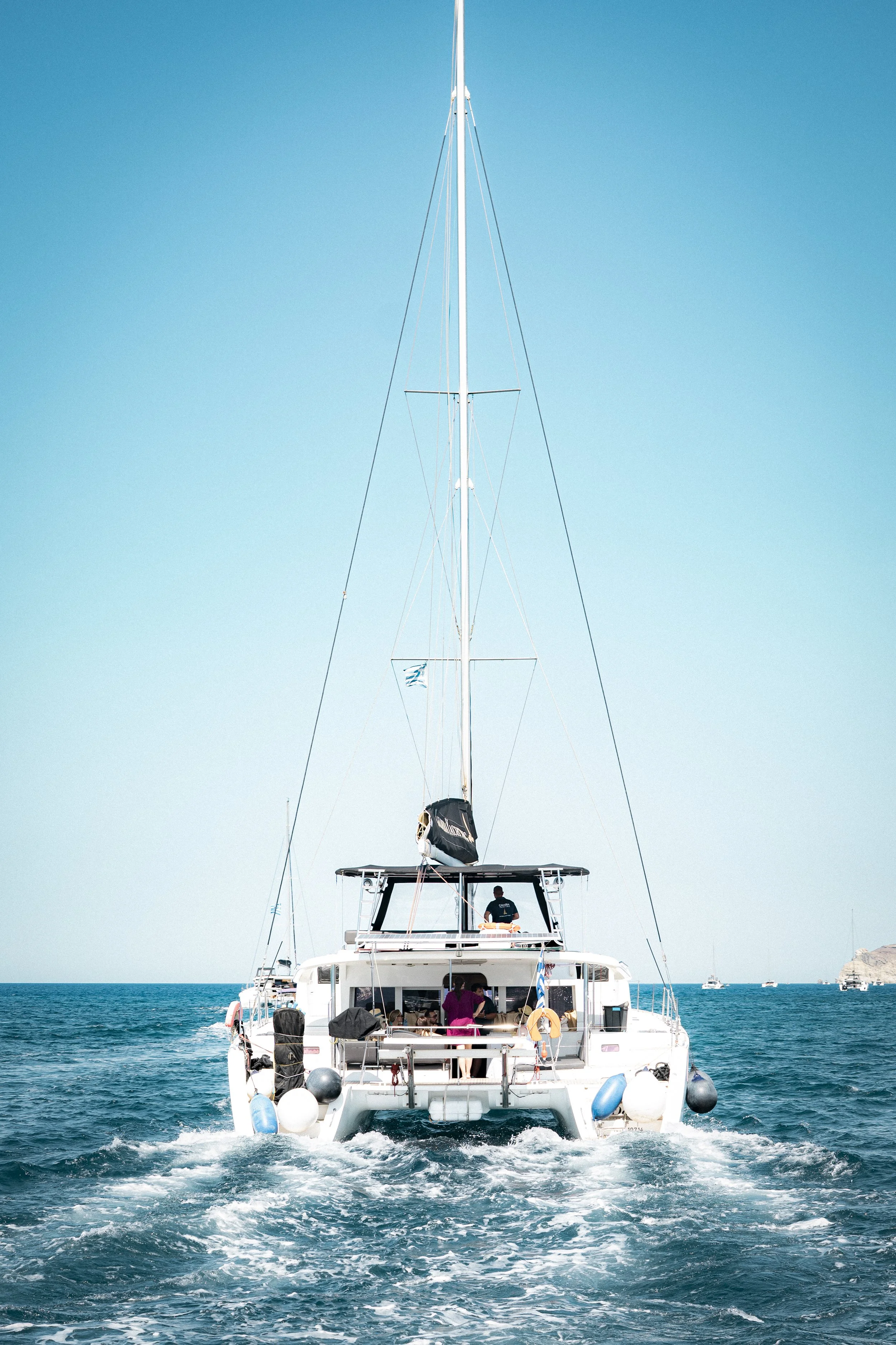 View of a white sailboat sailing on the ocean under a clear blue sky, with a few people on deck.