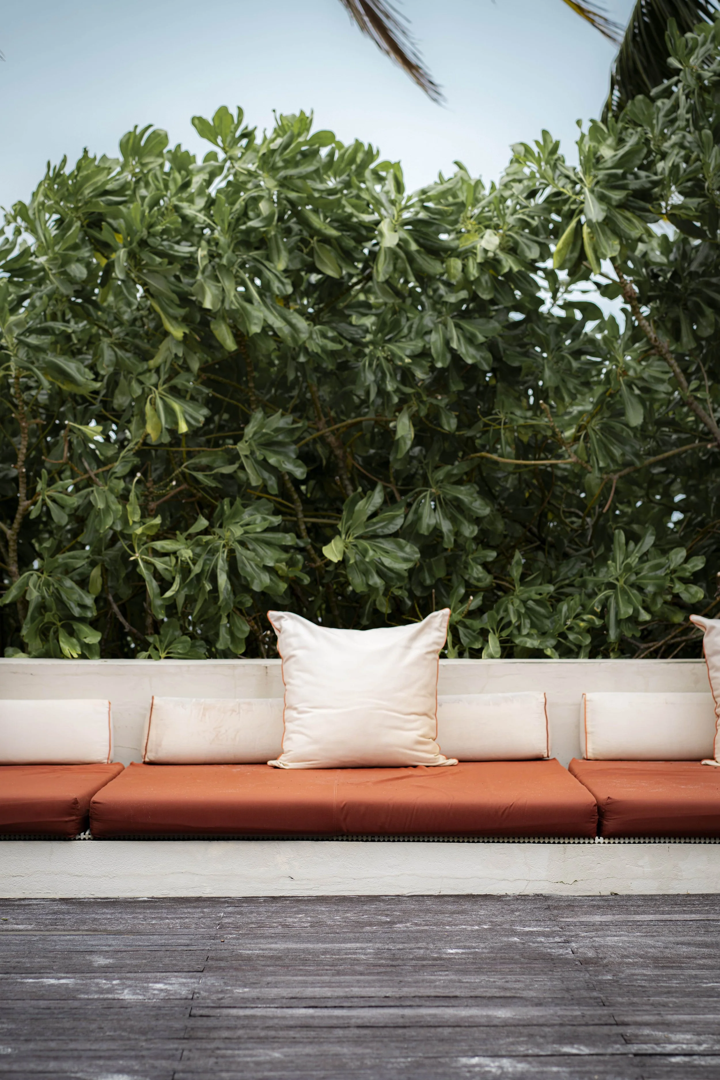 Outdoor seating area with a long orange cushion and several cream pillows, set against a background of lush green trees and a pale blue sky.