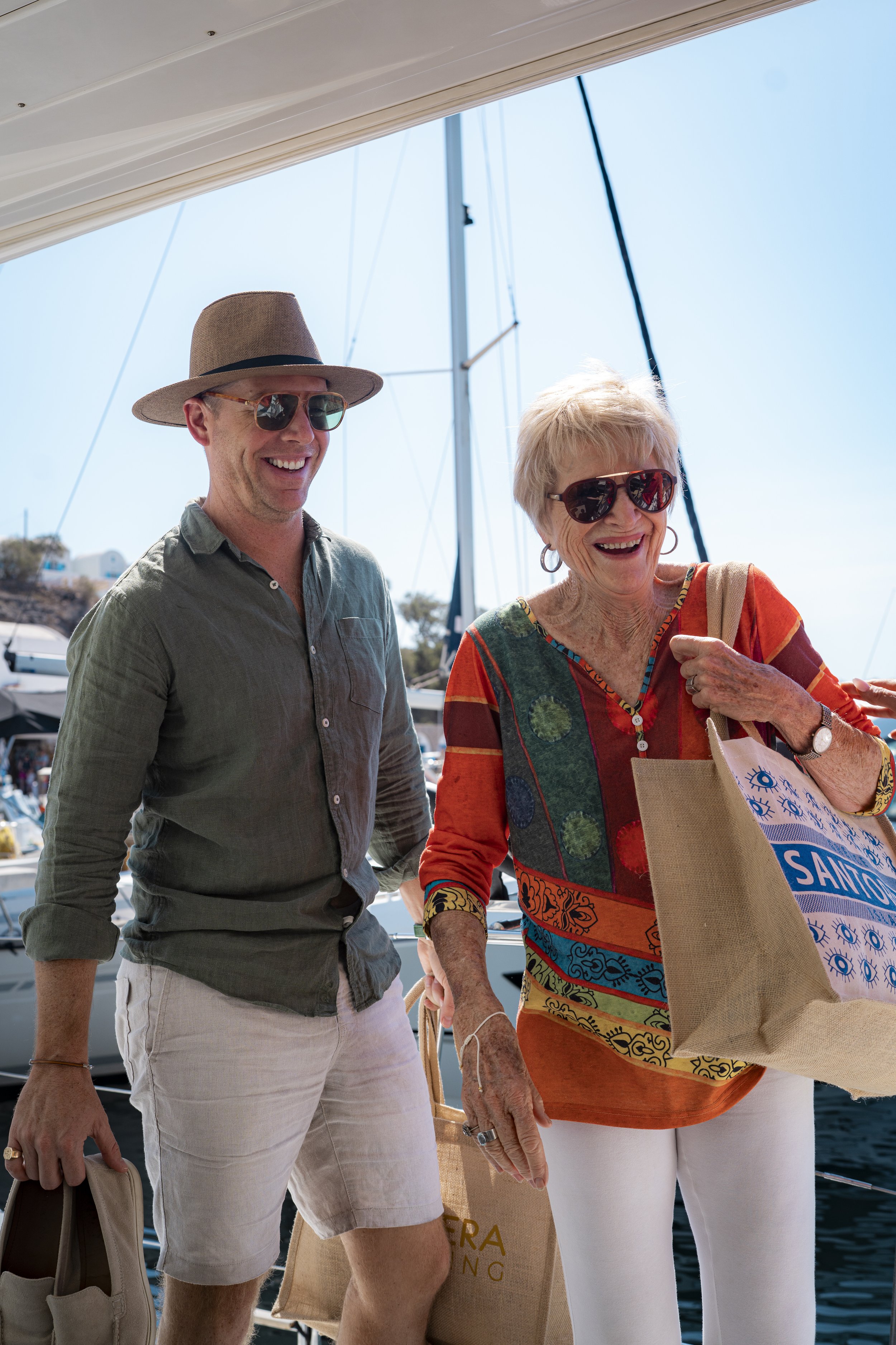 A man and an elderly woman smiling while at a marina, with boats and a clear sky in the background. They are wearing sunglasses and carrying shopping bags.