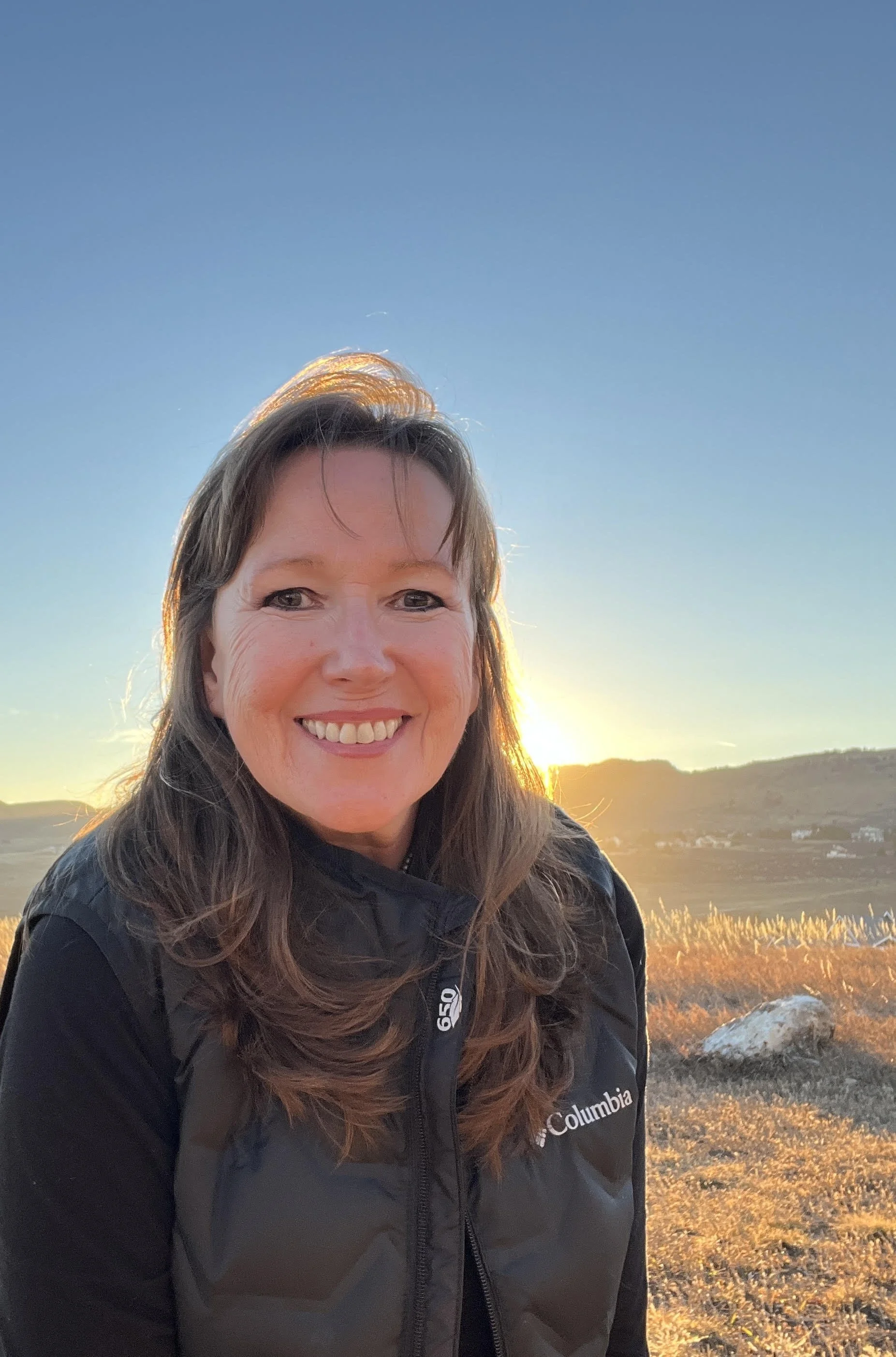 A woman with dark brown hair and bangs is smiling with a sunset in the background and blue sky