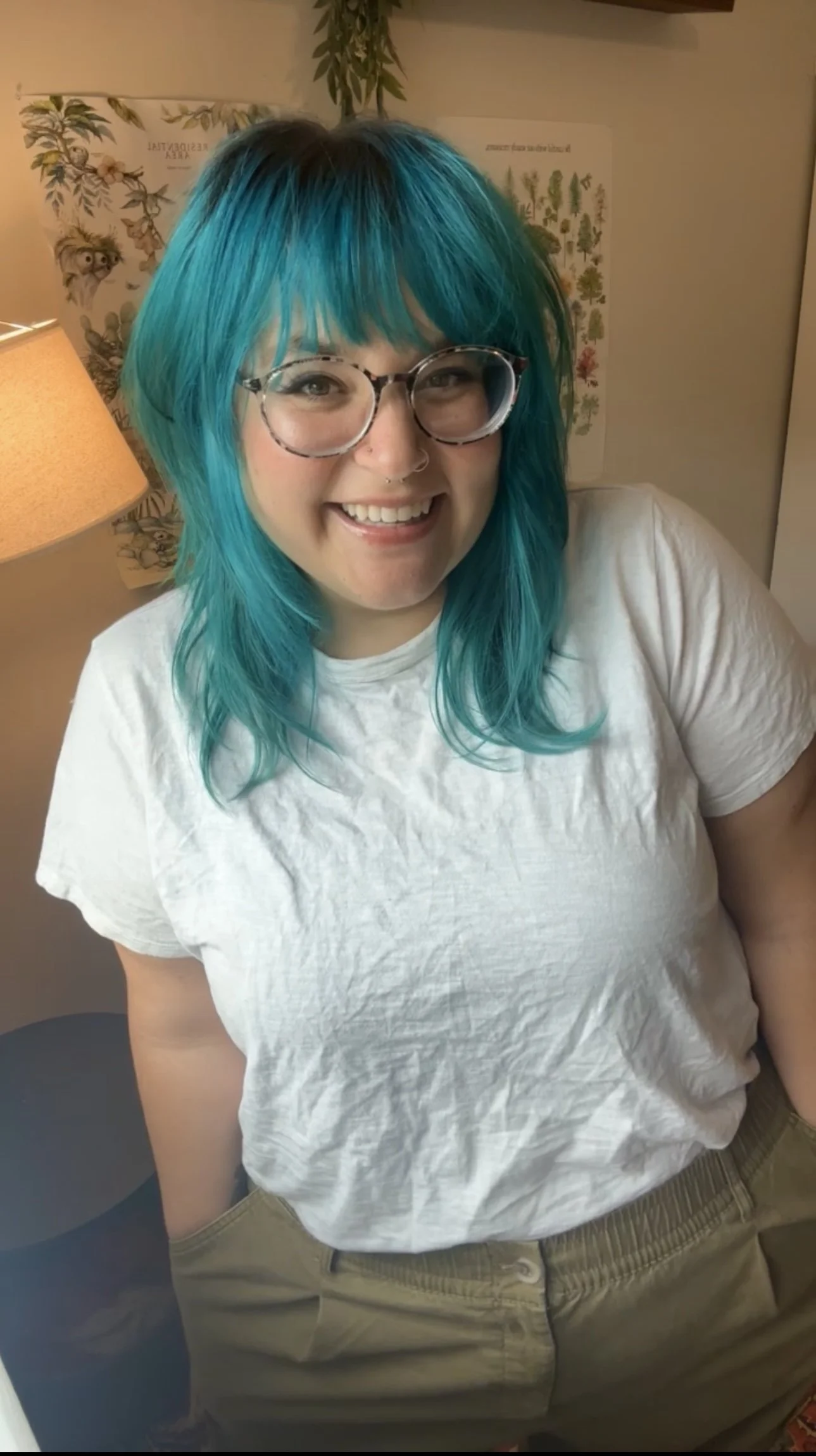 A smiling woman with teal-colored hair, wearing glasses and a white T-shirt, standing indoors with a lamp and botanical posters in the background.