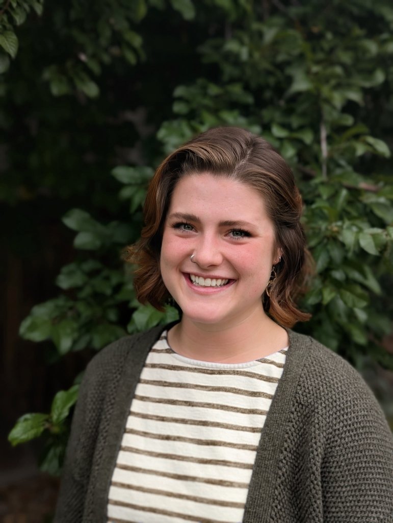A young woman with short wavy brown hair, wearing a nose ring, a striped t-shirt, and a dark cardigan, smiling outdoors with green foliage in the background.