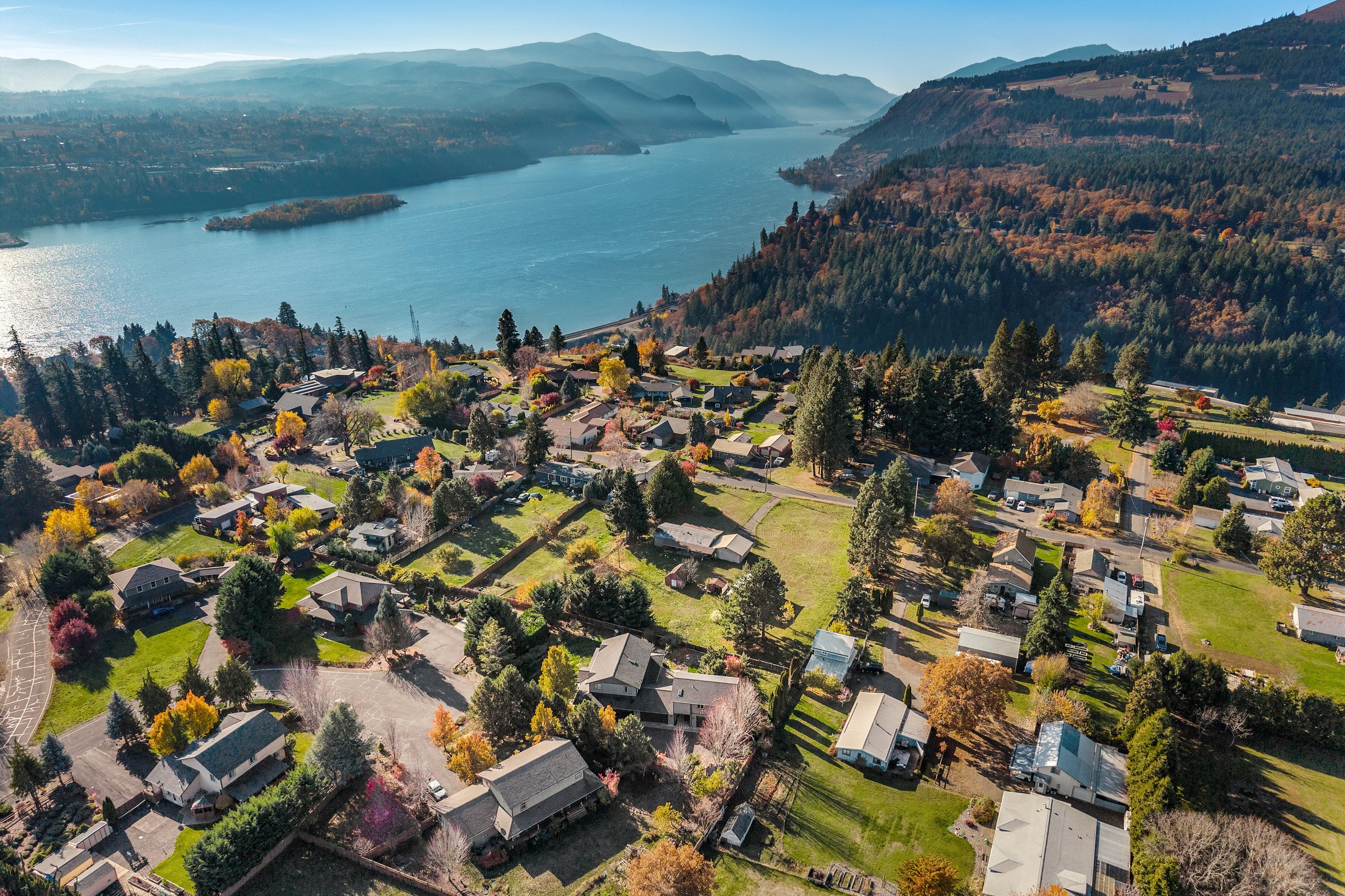 An aerial view of a lakeside residential neighborhood with houses, trees, and roads surrounded by mountains and a large body of water.