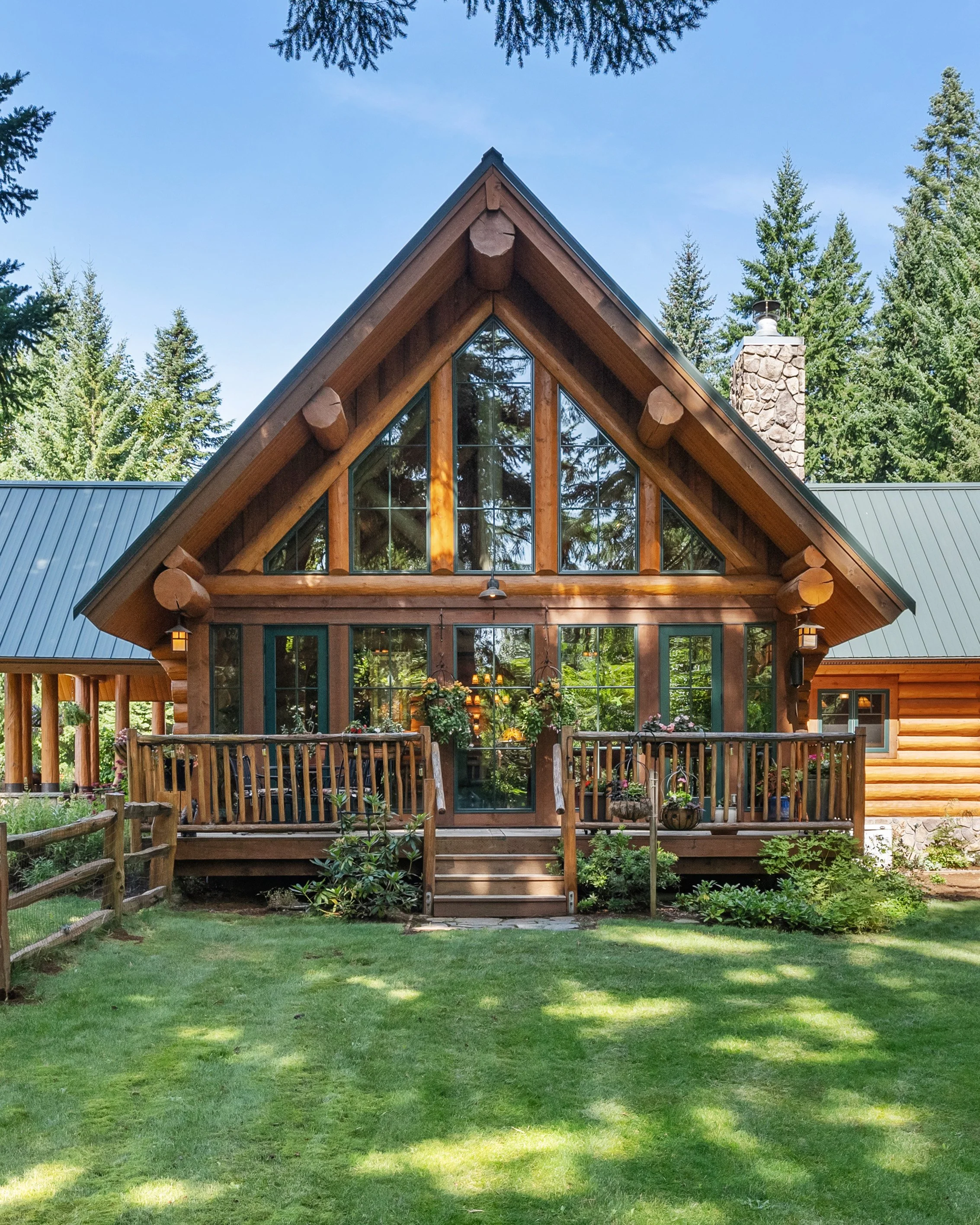 A rustic log cabin with large triangular windows and a green metal roof, surrounded by tall pine trees and a well-maintained grassy yard.