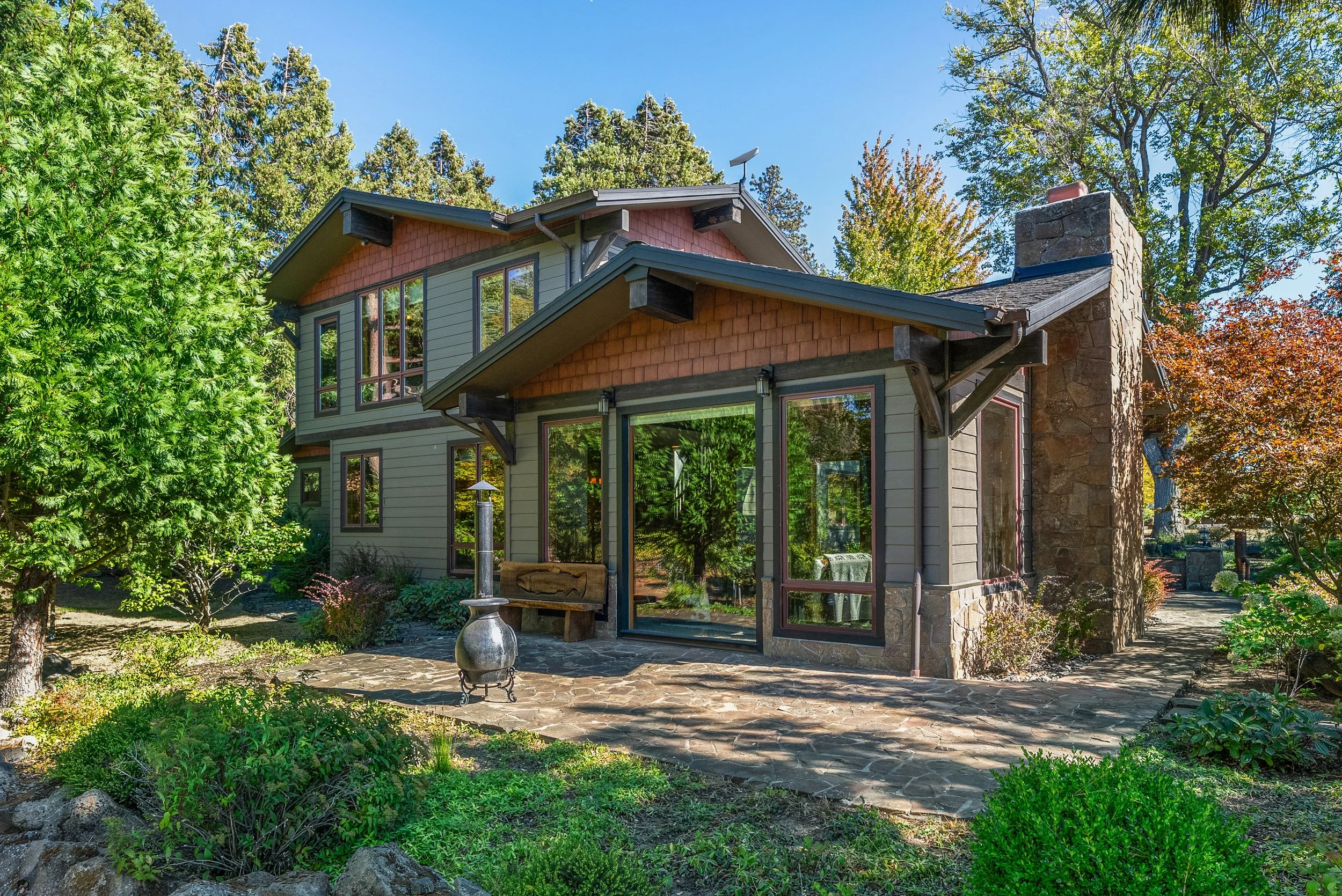 A two-story house with a stone chimney, large glass sliding door, surrounded by green trees and shrubs, with a stone patio in the foreground.