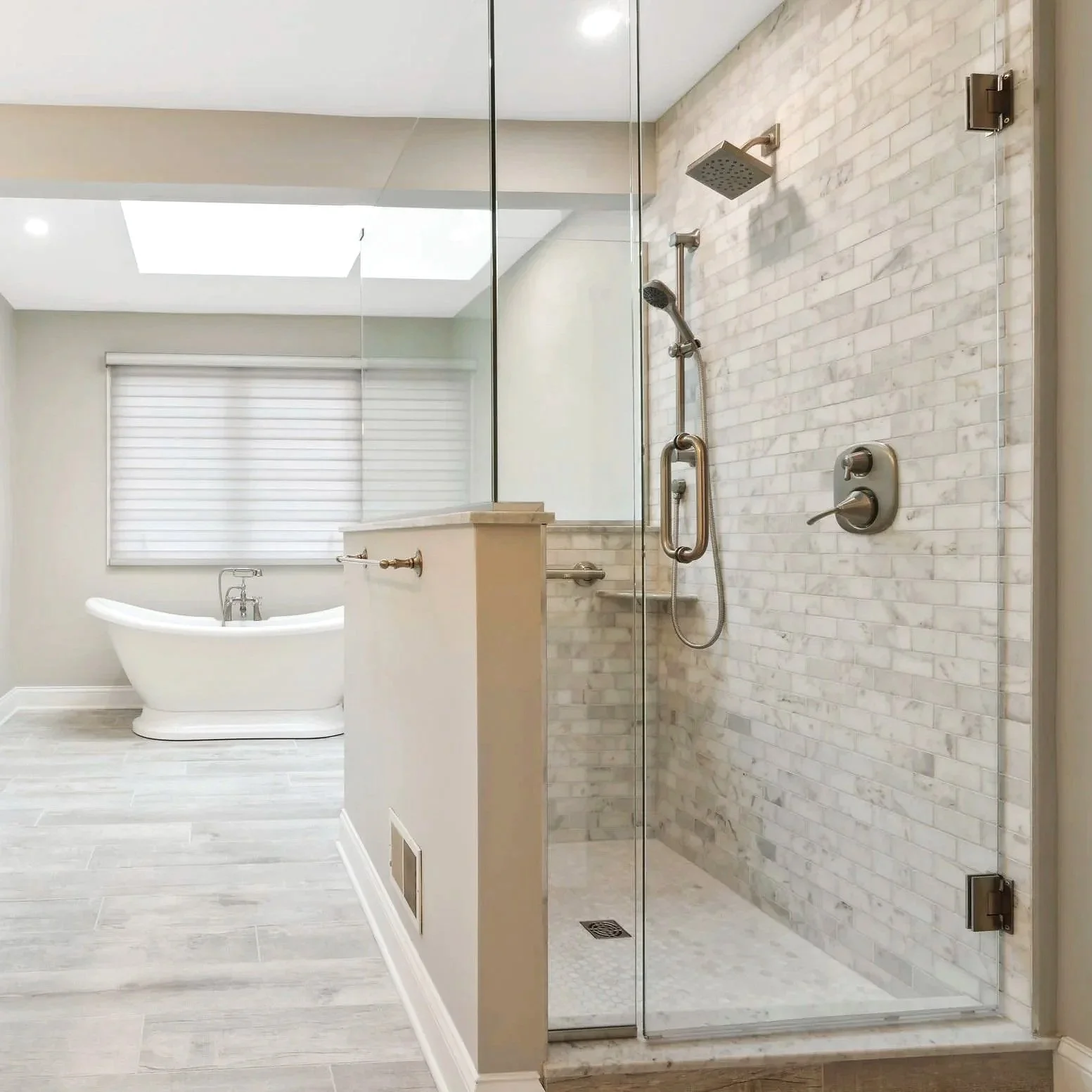 Bathroom with a glass shower enclosure on the right featuring marble tiles, a showerhead, and controls, and a freestanding bathtub in the background, with a window and blinds on the far wall.
