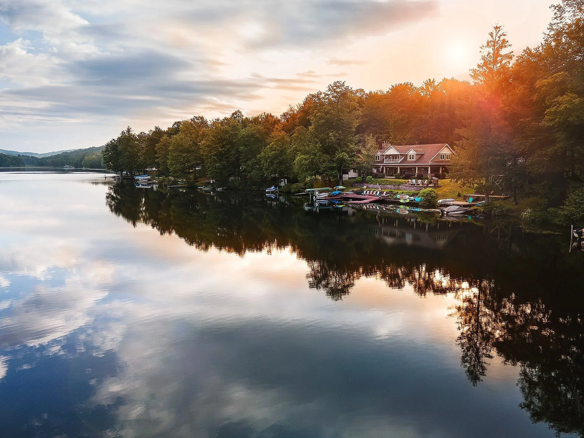 Sunset over a calm lake with trees and houses along the shoreline, and boats docked by the water.