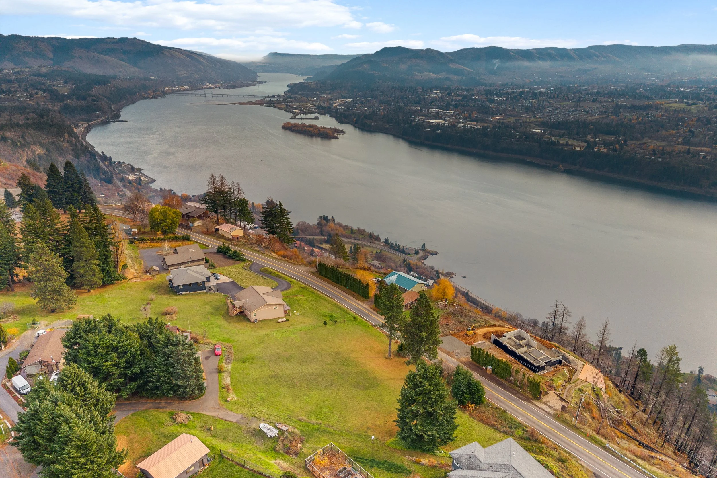 Aerial view of a residential area along a river with a bridge, surrounded by hills and trees, some areas under construction, in a mountainous landscape.