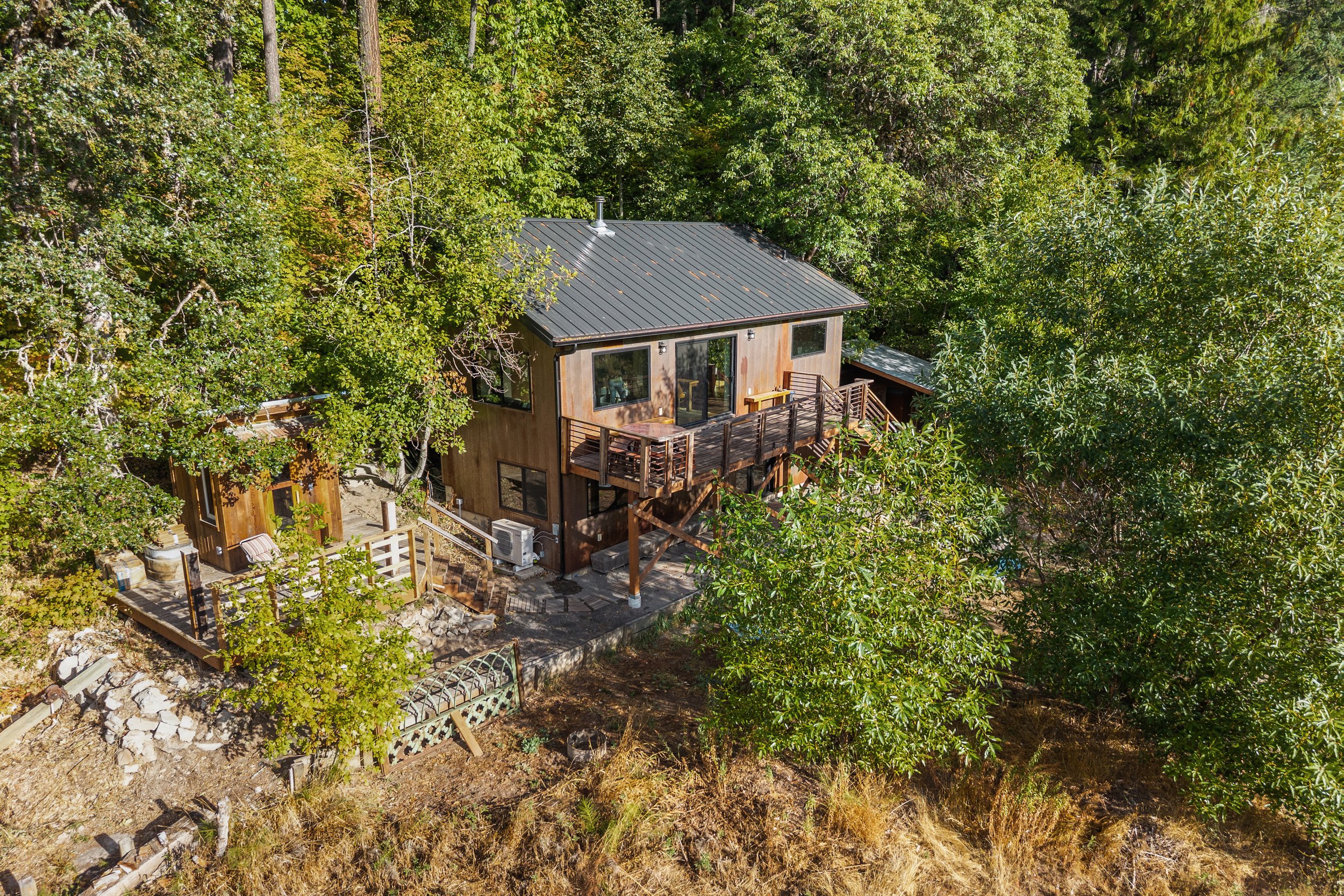 An aerial view of a small wooden house with a metal roof situated among green trees. The house has a balcony and a patio area with outdoor furniture.