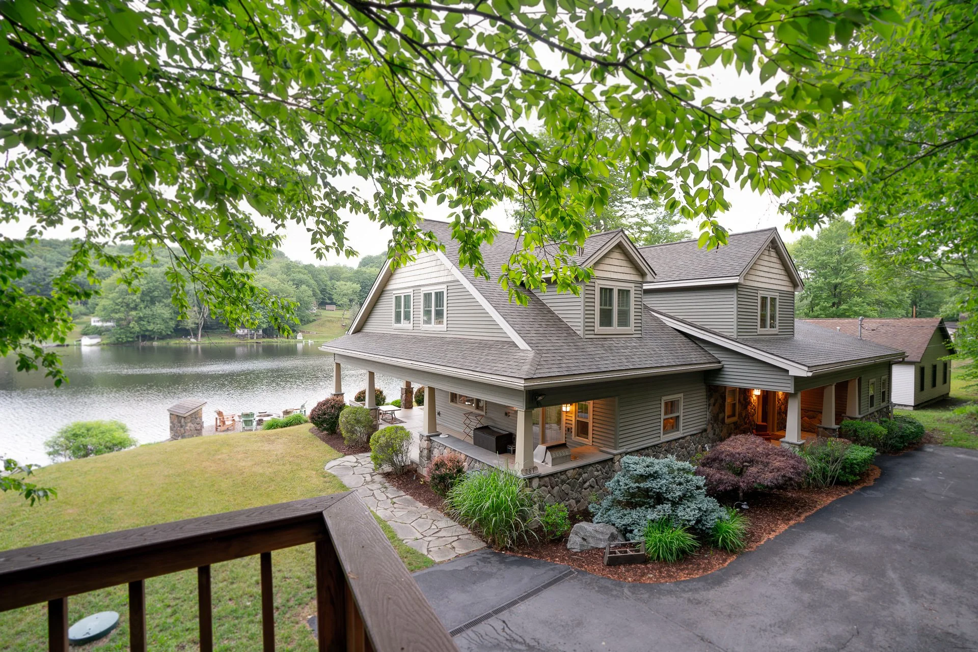 Two-story house with gray siding and a stone foundation, nestled beside a lake, with a deck and landscaped garden in the foreground and a wooded area across the lake.
