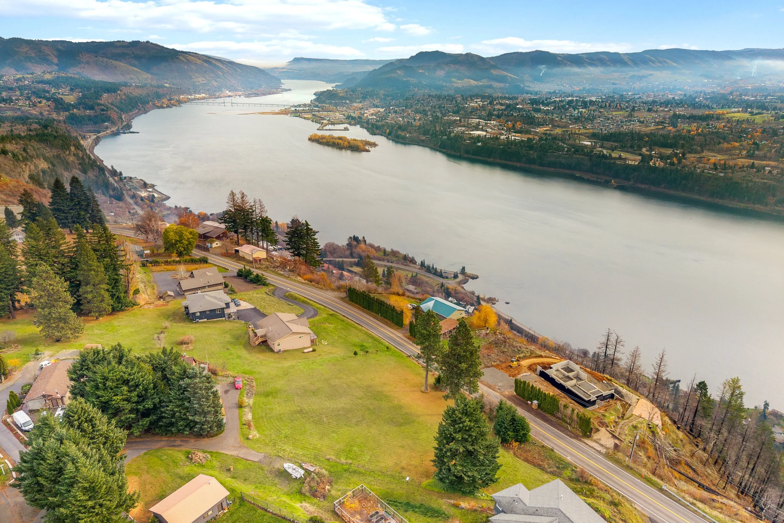 Aerial view of a riverside neighborhood with houses, trees, and a large body of water, surrounded by hills and mountains.