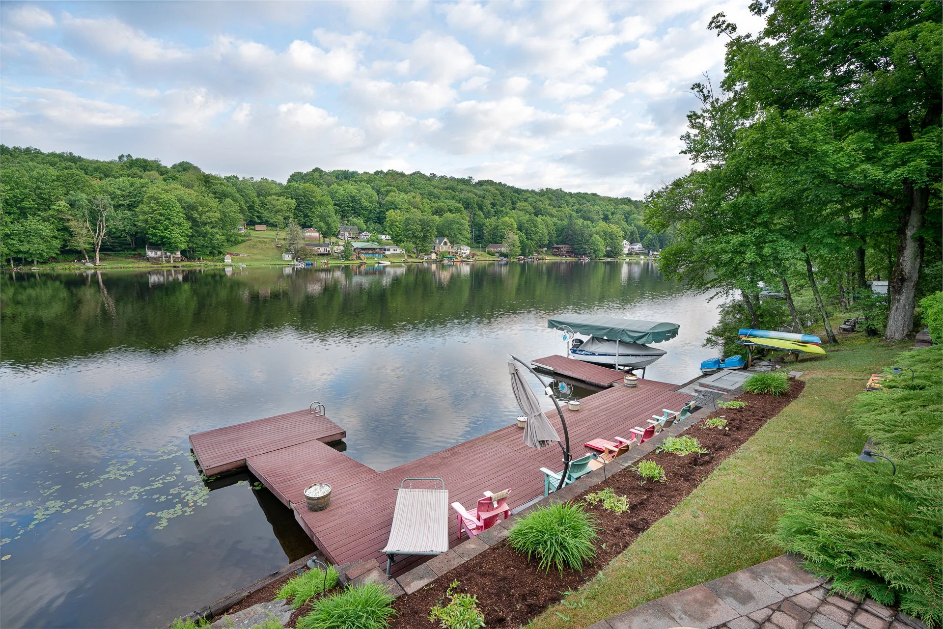 A peaceful lakeside scene featuring a dock with colorful lawn chairs, umbrellas, and kayaks on the grassy shore. Calm water reflects surrounding green trees and a partly cloudy sky.
