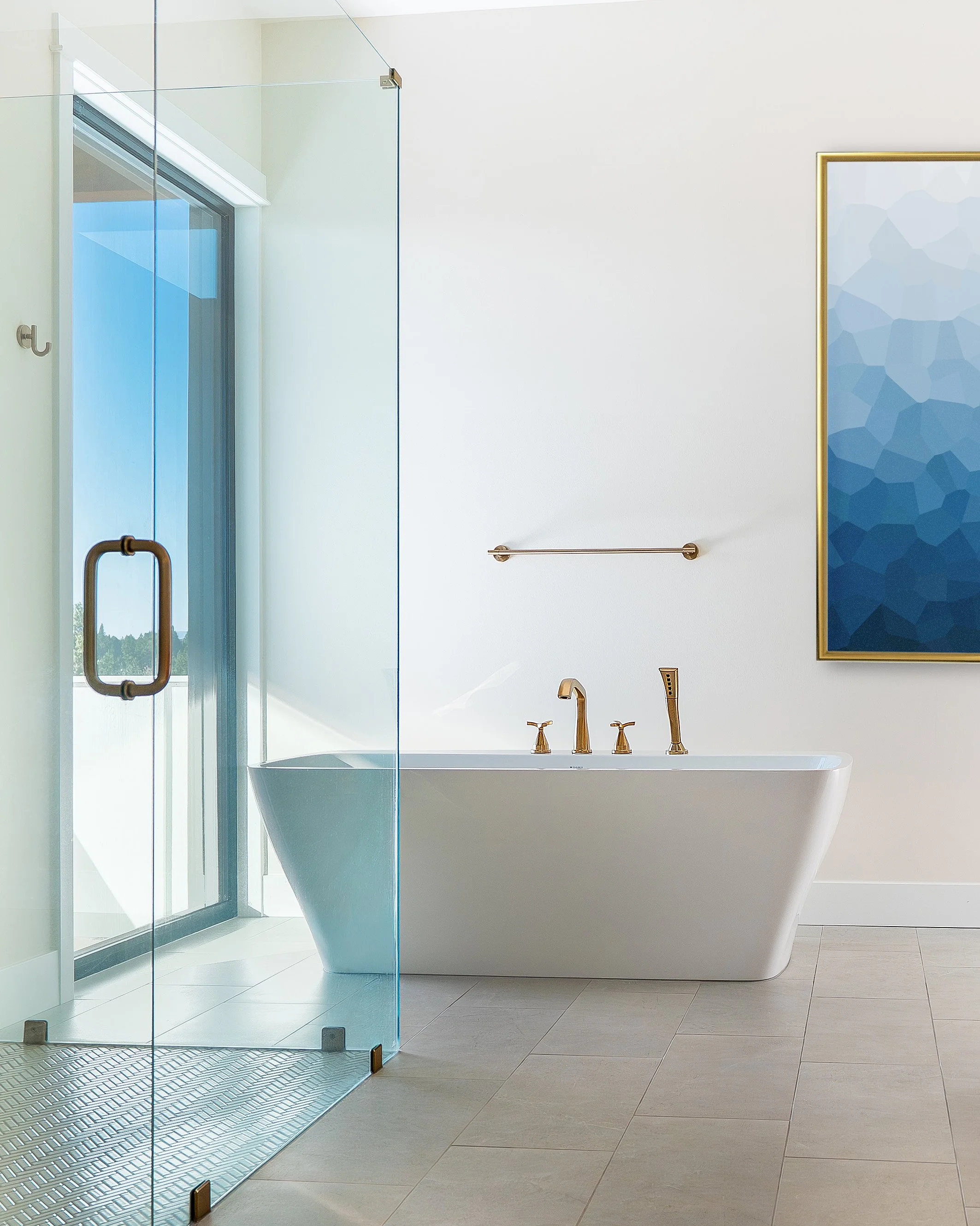 Modern bathroom with glass-enclosed shower, white bathtub with gold fixtures, and abstract blue artwork on white wall.
