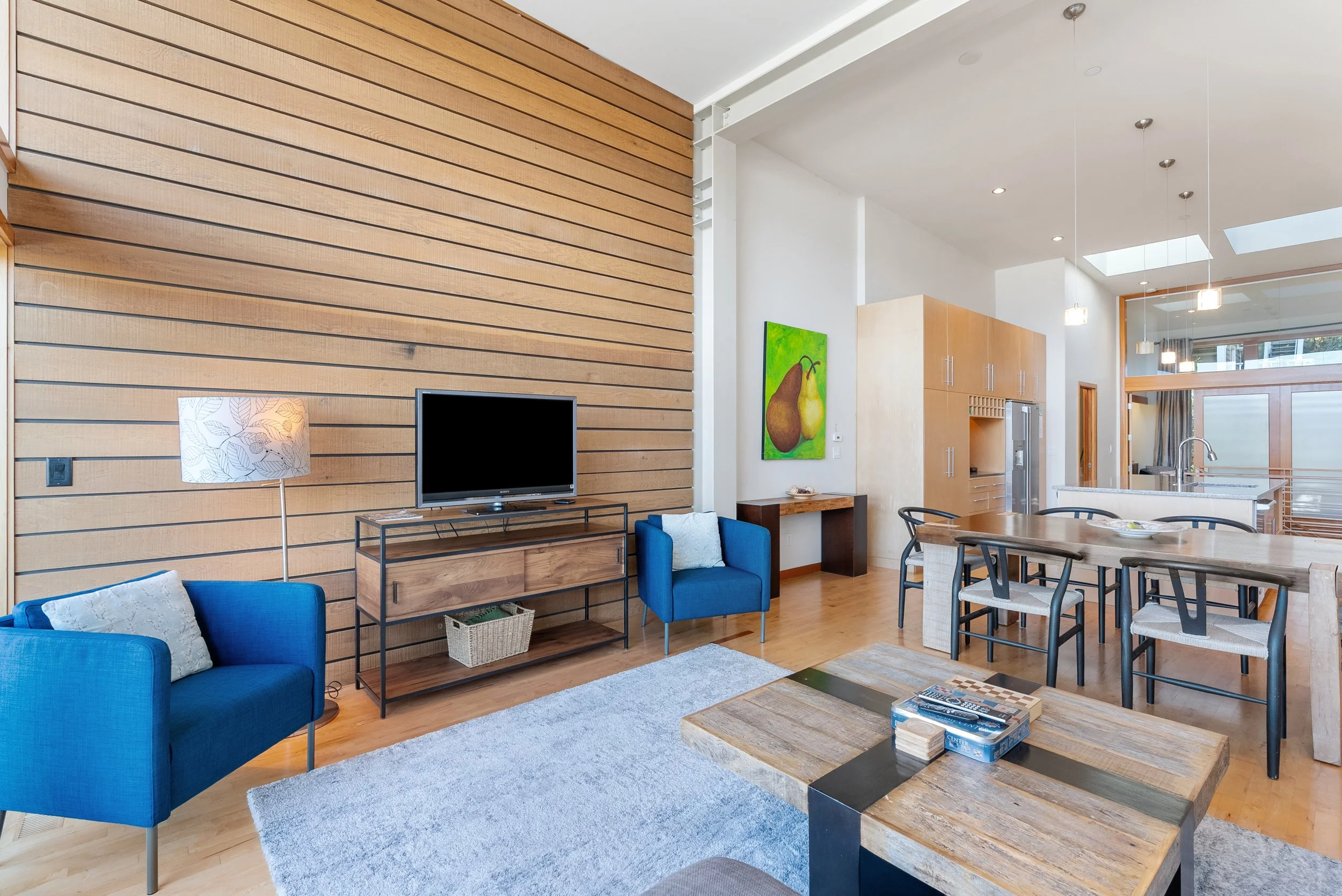Living room with wooden wall, blue armchair, flat-screen TV, modern chandelier, dining area with black chairs, kitchen in the background, artwork of pears on the wall, and wooden furniture.