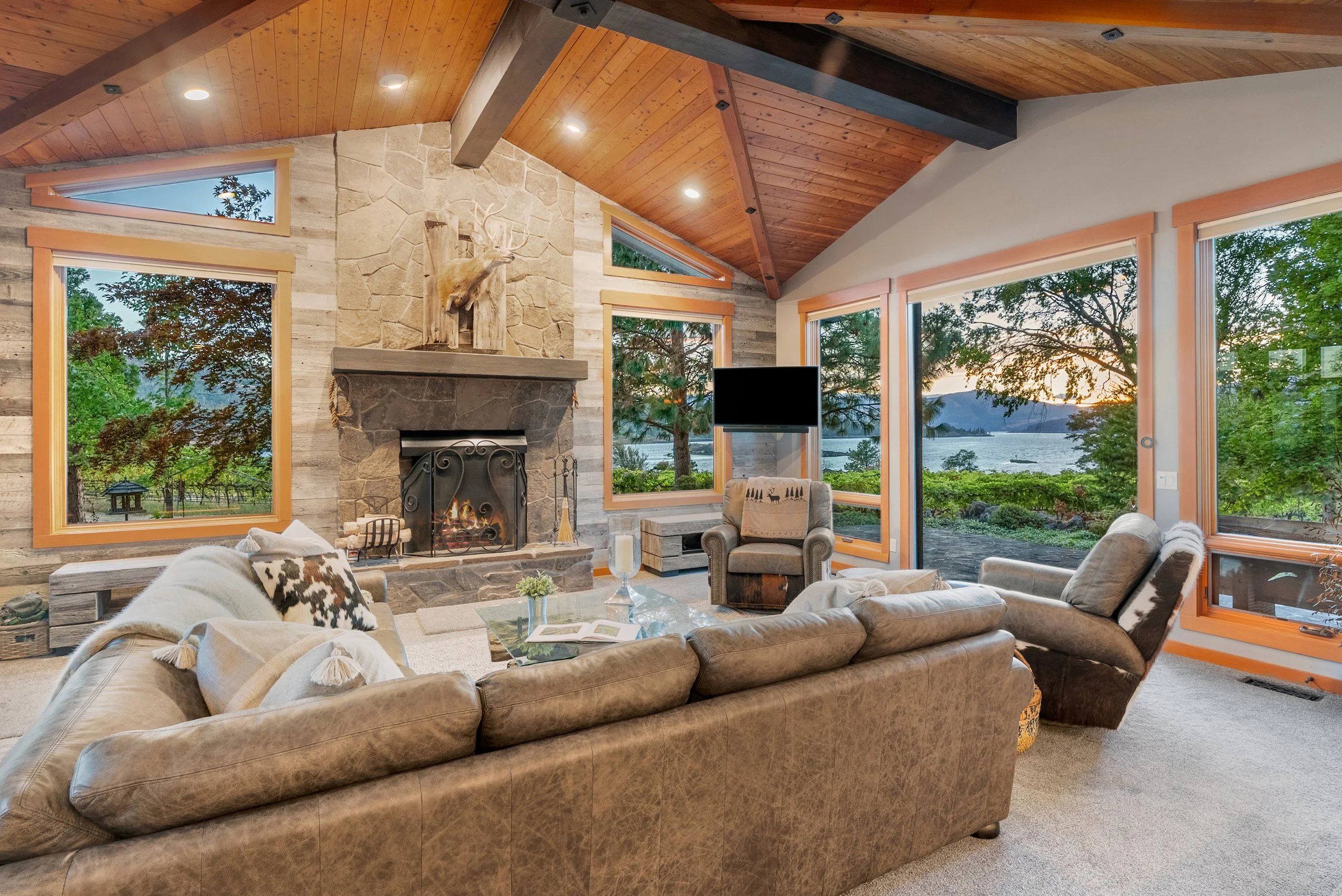 Living room with a large sectional sofa, a stone fireplace, and multiple windows showing a scenic lake view at sunset.