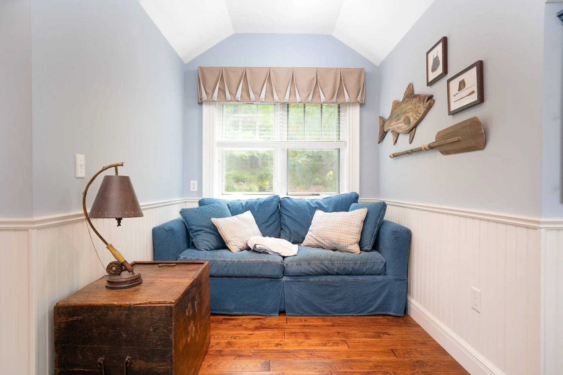 A cozy living room with a blue sofa, beige checkered pillows, wooden side table with a vintage lamp, and nautical-themed wall decorations, including a fish and oars.