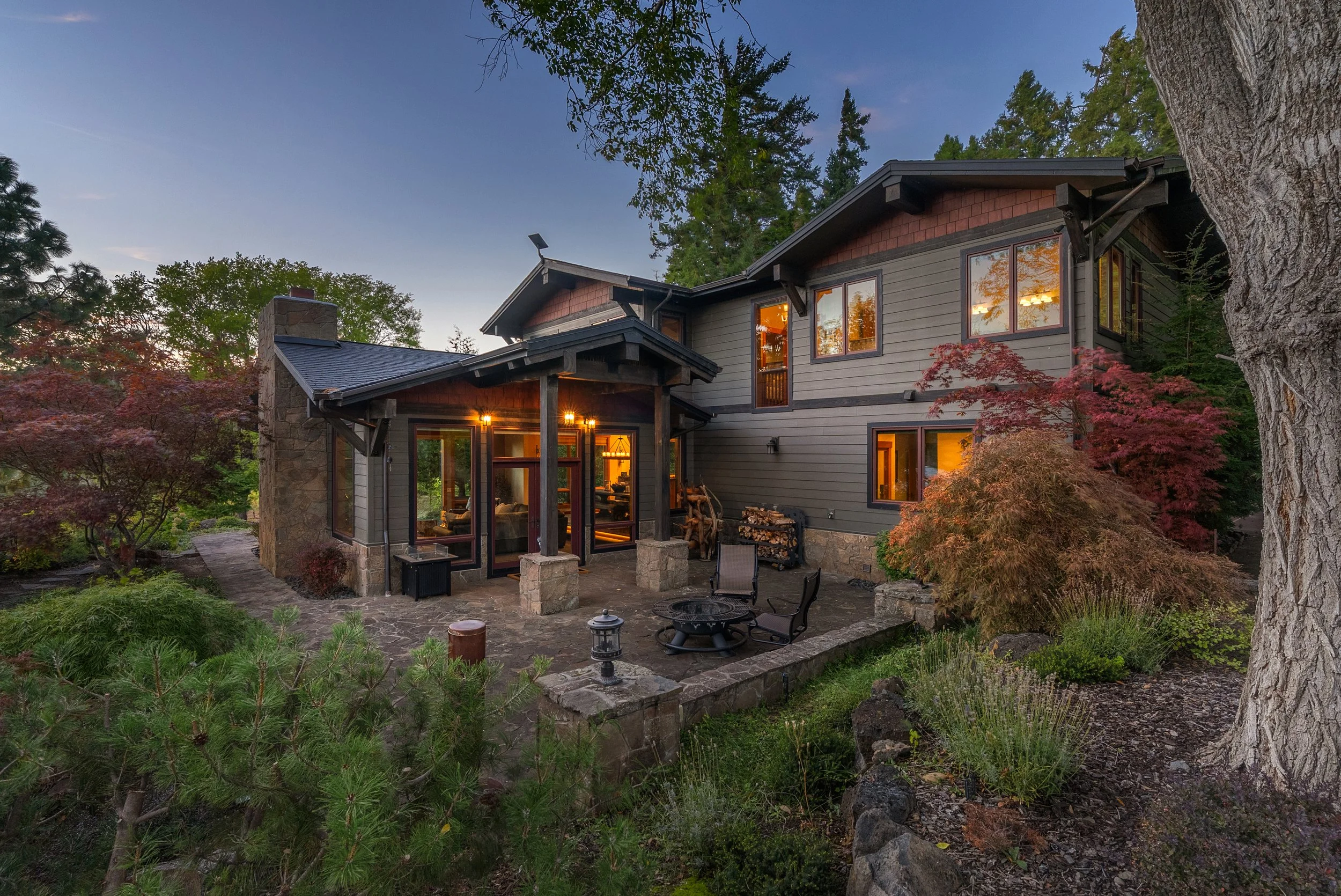 Exterior view of a two-story house with a stone patio, surrounded by trees and landscaping, at dusk with interior lights on.