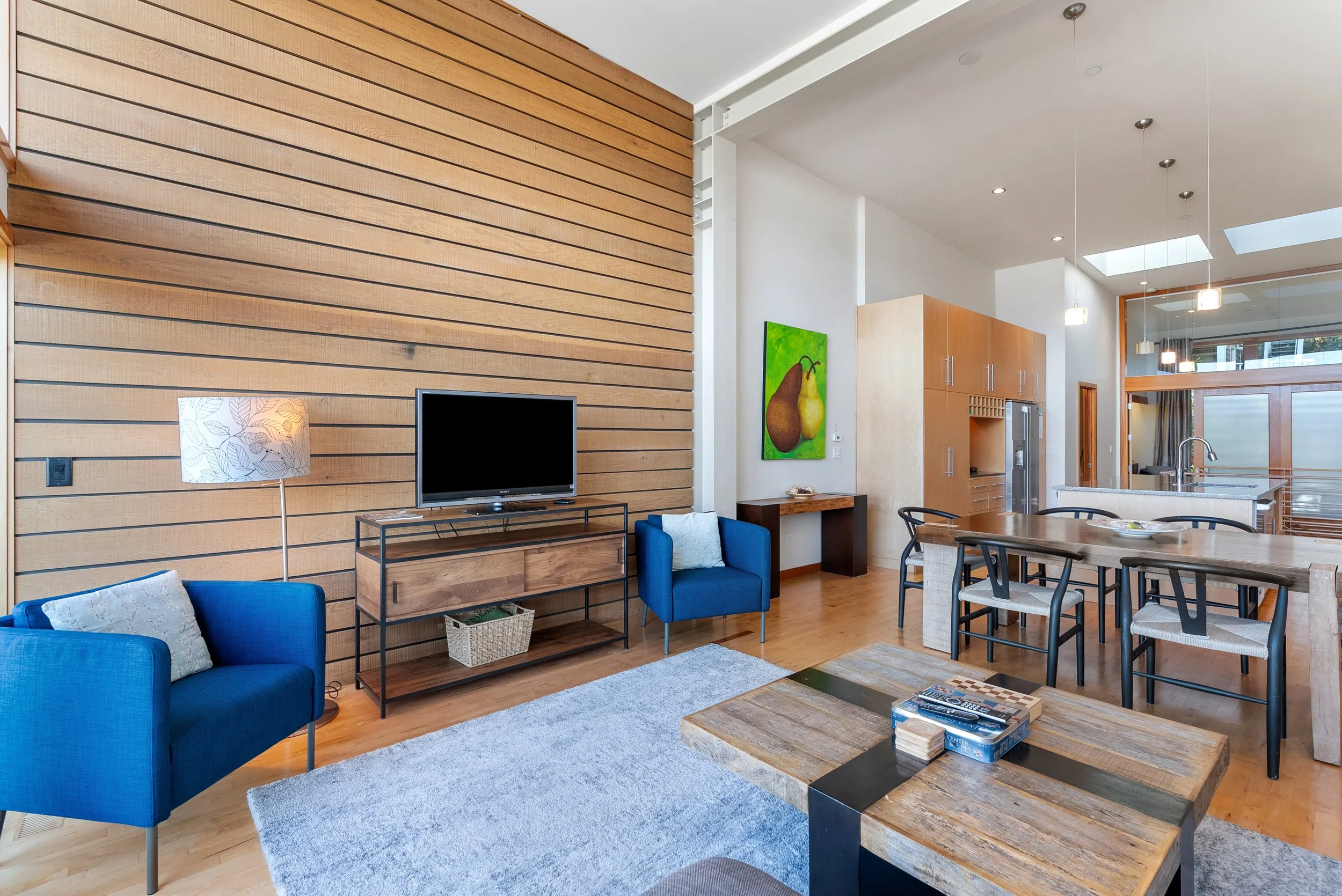A modern living room with a blue armchair, a wooden coffee table with board games, a flat-screen TV on a stand, a floor lamp, and a light wood-paneled accent wall. The room opens to a kitchen and dining area with a wooden table and chairs, stainless 