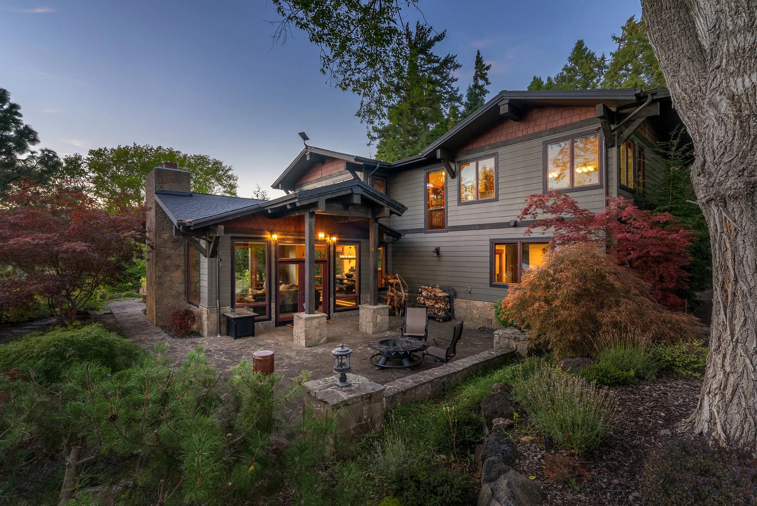 A two-story house with large windows, surrounded by trees and garden, illuminated at dusk with outdoor patio furniture and wood logs nearby.
