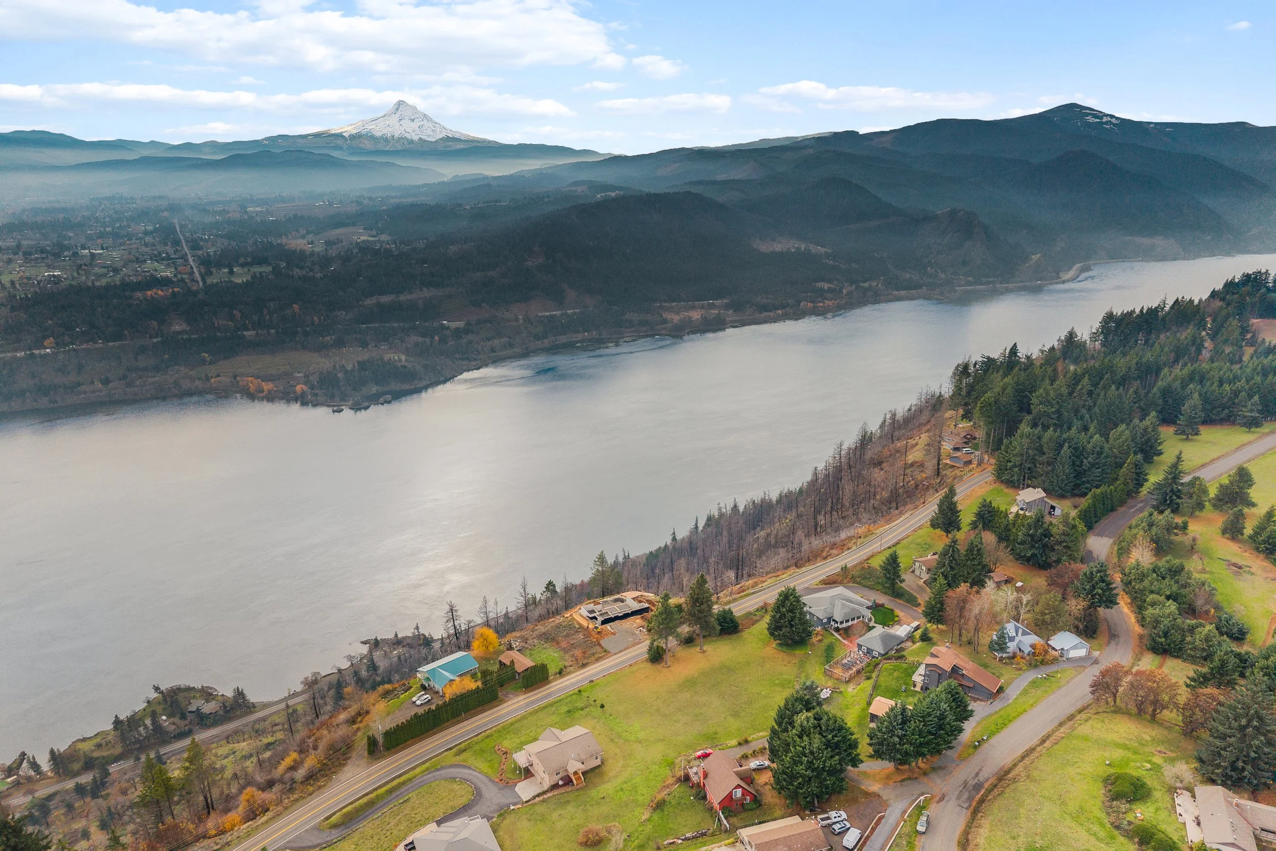 Aerial view of a river with a small residential neighborhood along its edge, surrounded by green trees, hills, and mountains in the background, under a partly cloudy sky.