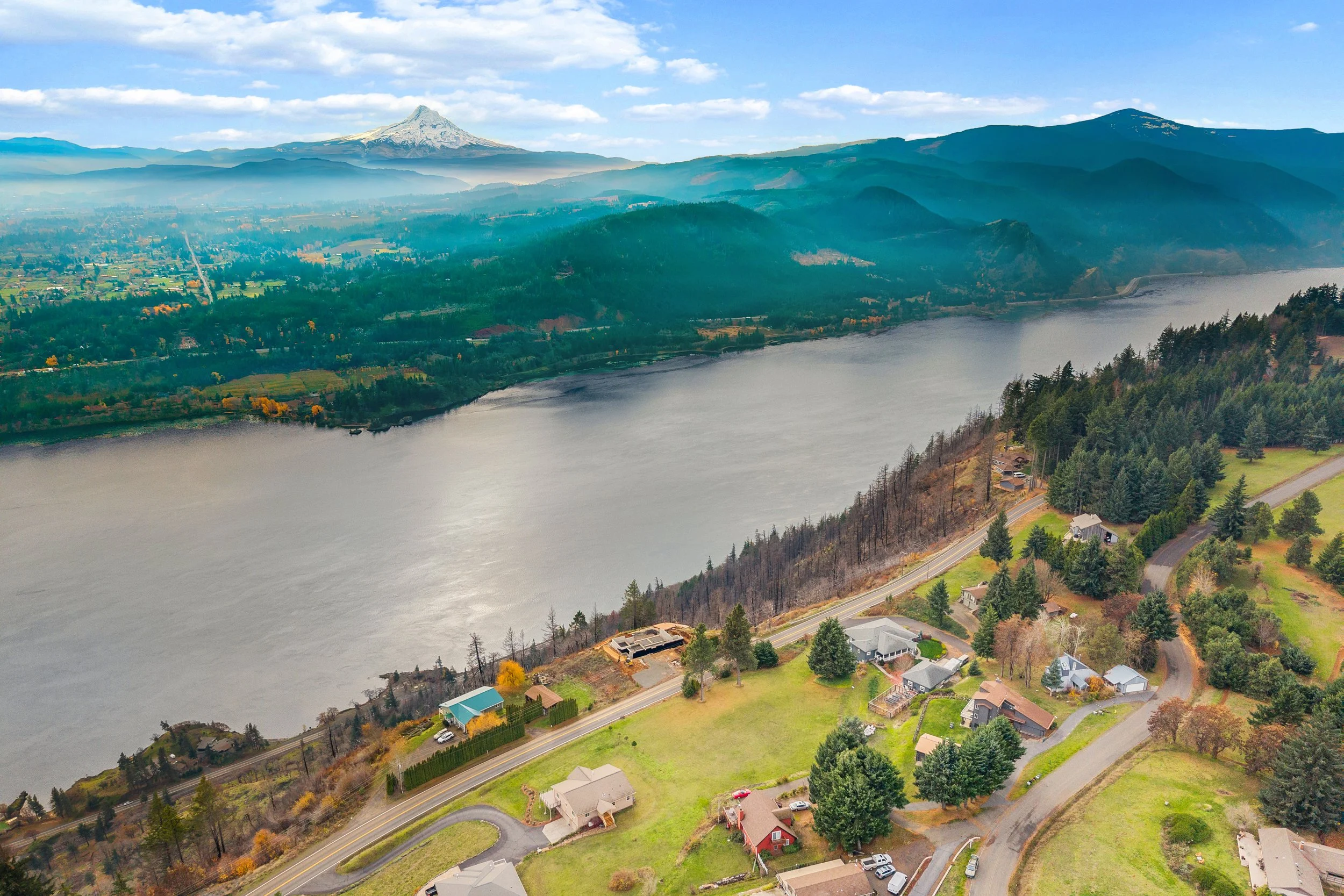 Aerial view of a river with residential houses along the bank, surrounded by green trees and rolling hills, with snow-capped mountains in the distance under a partly cloudy sky.