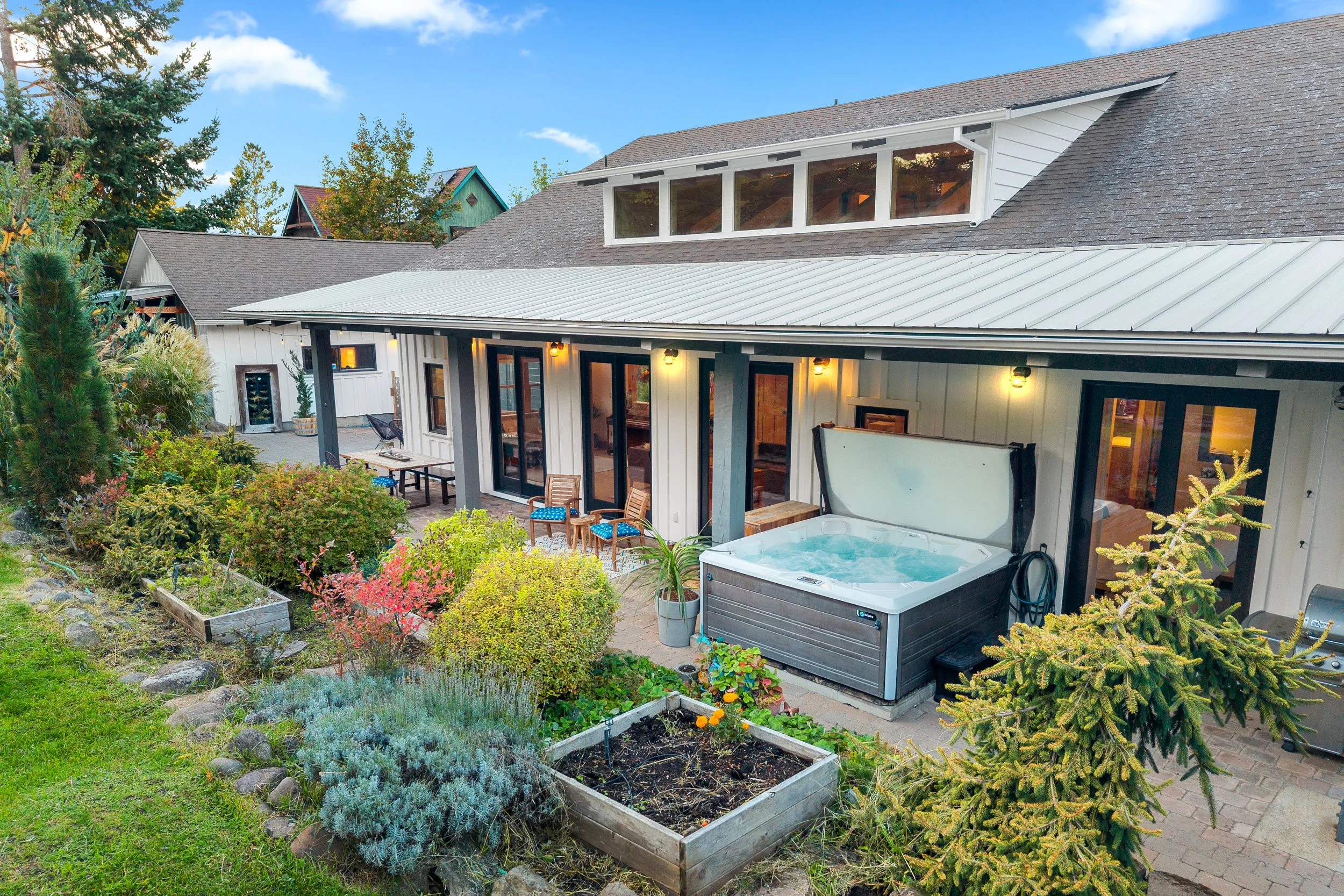 A backyard patio with a hot tub, outdoor seating, and a garden with plants and flowers adjacent to a house with large windows and a metal roof.