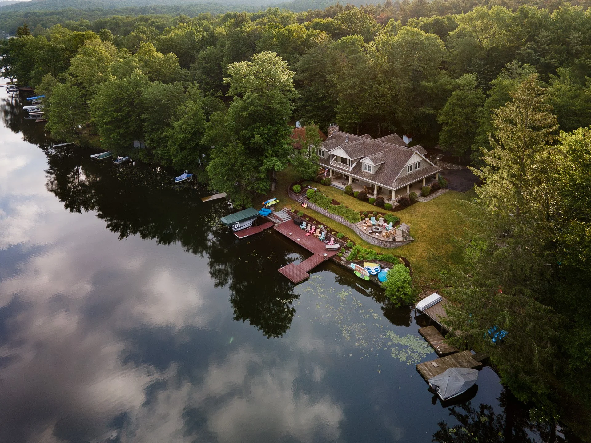 A lakefront house with a backyard patio, surrounded by trees. The area features lounge chairs, a fire pit, and a dock with boats and watercraft. The reflection of the sky and clouds can be seen in the water.