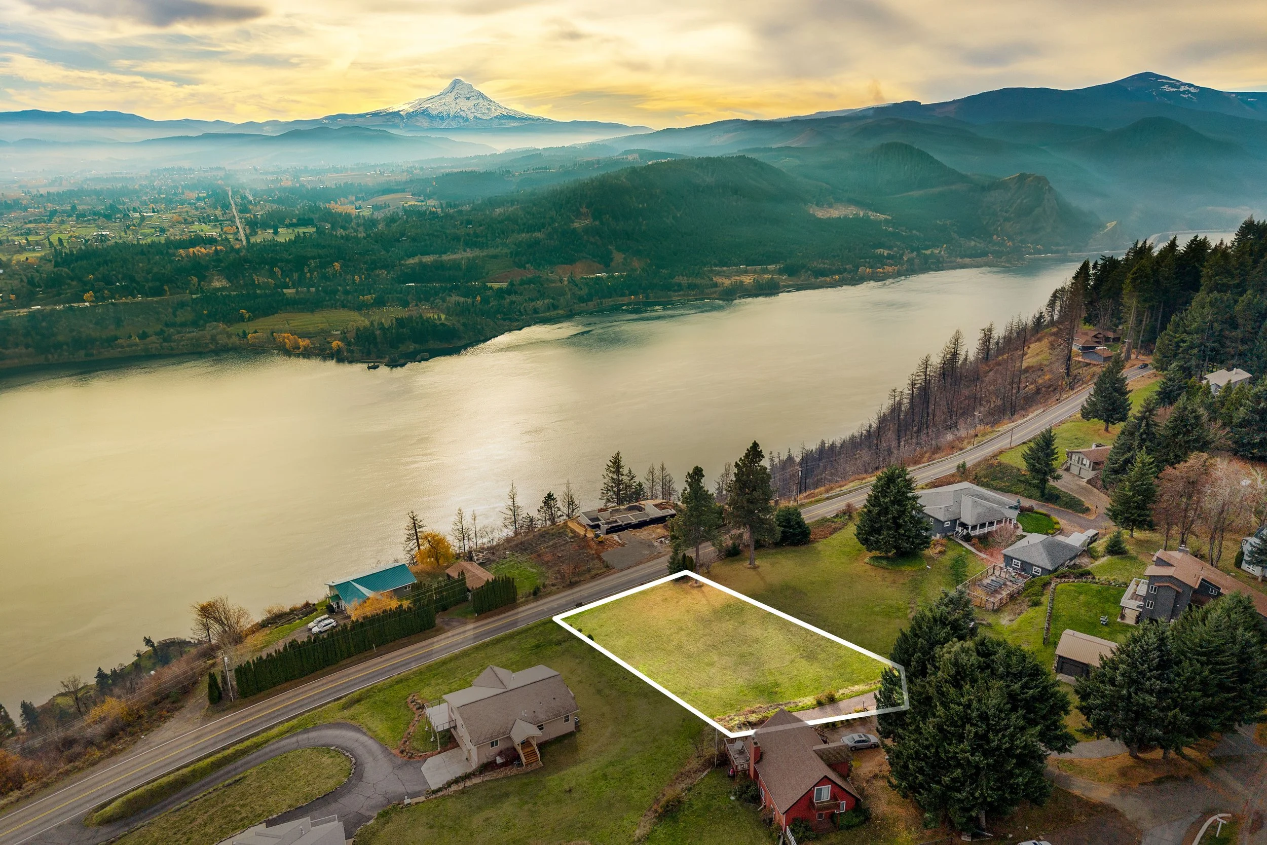 A plot of land outlined in white, located near a river and surrounded by houses, trees, and a road, with a mountainous landscape and city in the distance.