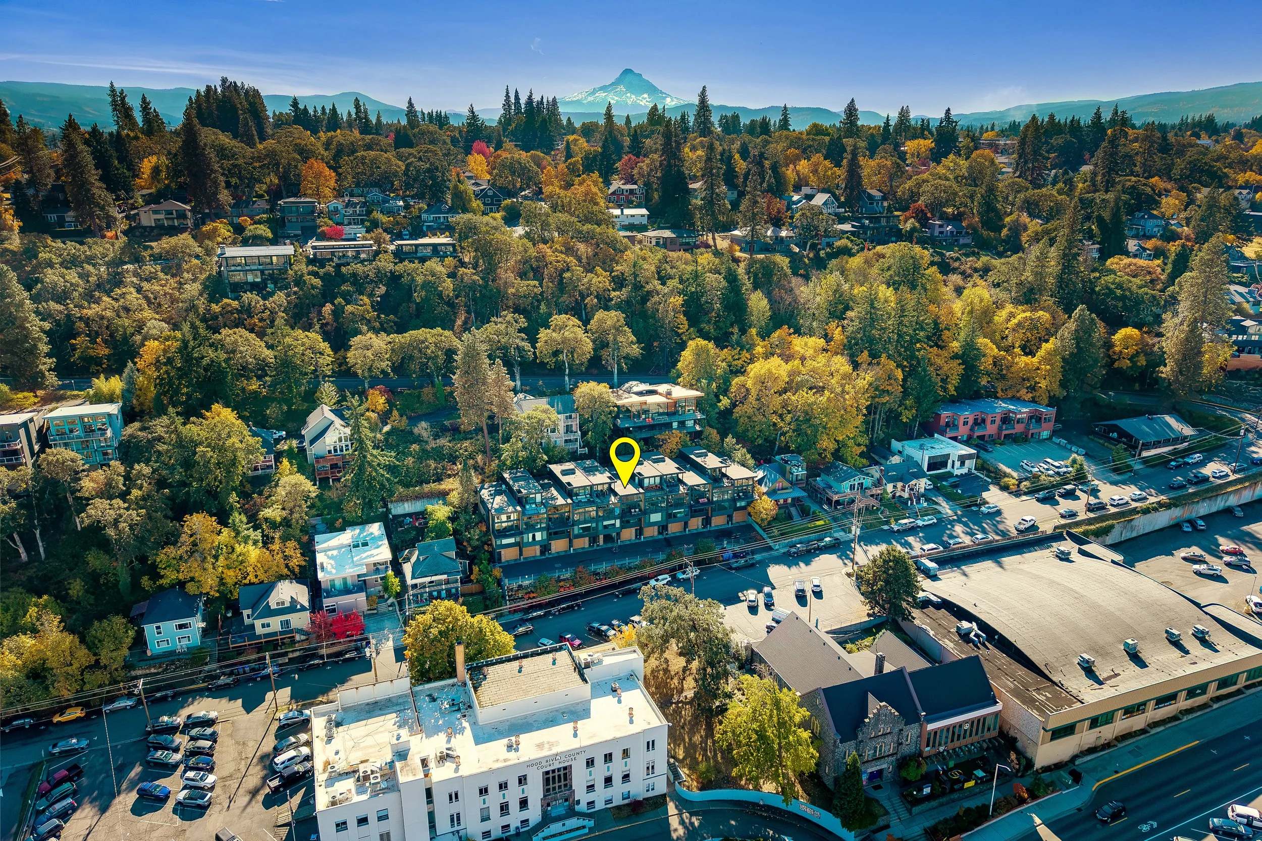 Aerial view of a hilly town with residential houses, trees with autumn foliage, a parking lot, and distant mountains with a snow-capped peak.