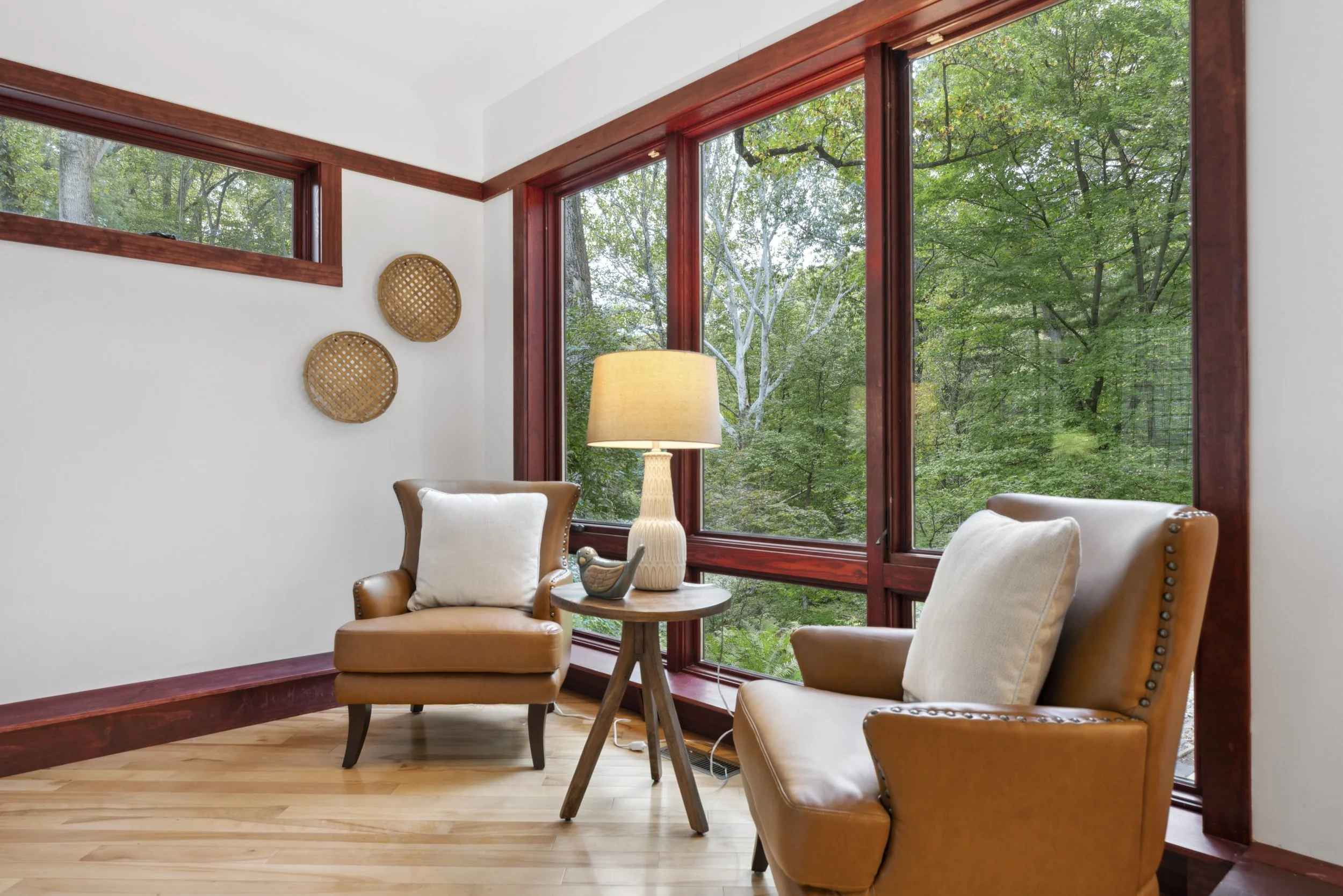 Cozy sitting area with two tan armchairs with pillows, a small wooden side table with a lamp and decorative bird, large windows with a view of green trees outside, and wall decor of two round wicker trays.