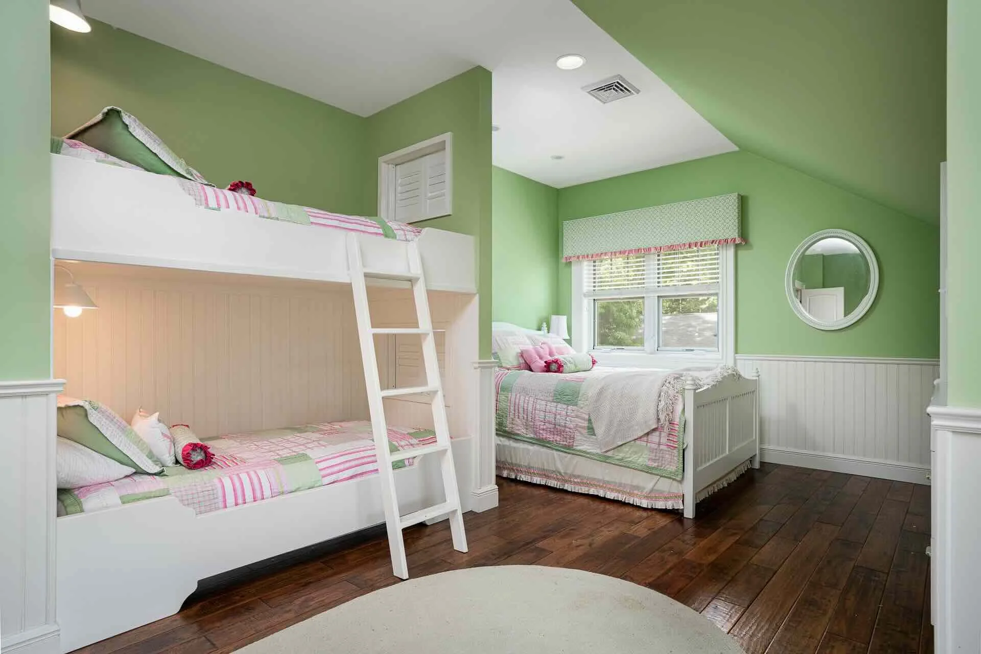 A bedroom with green walls and white furniture featuring a bunk bed with pink and green bedding, and a separate bed near a window with matching bedding and pillows. The room has hardwood floors, a circular mirror on the wall, and a window with blinds
