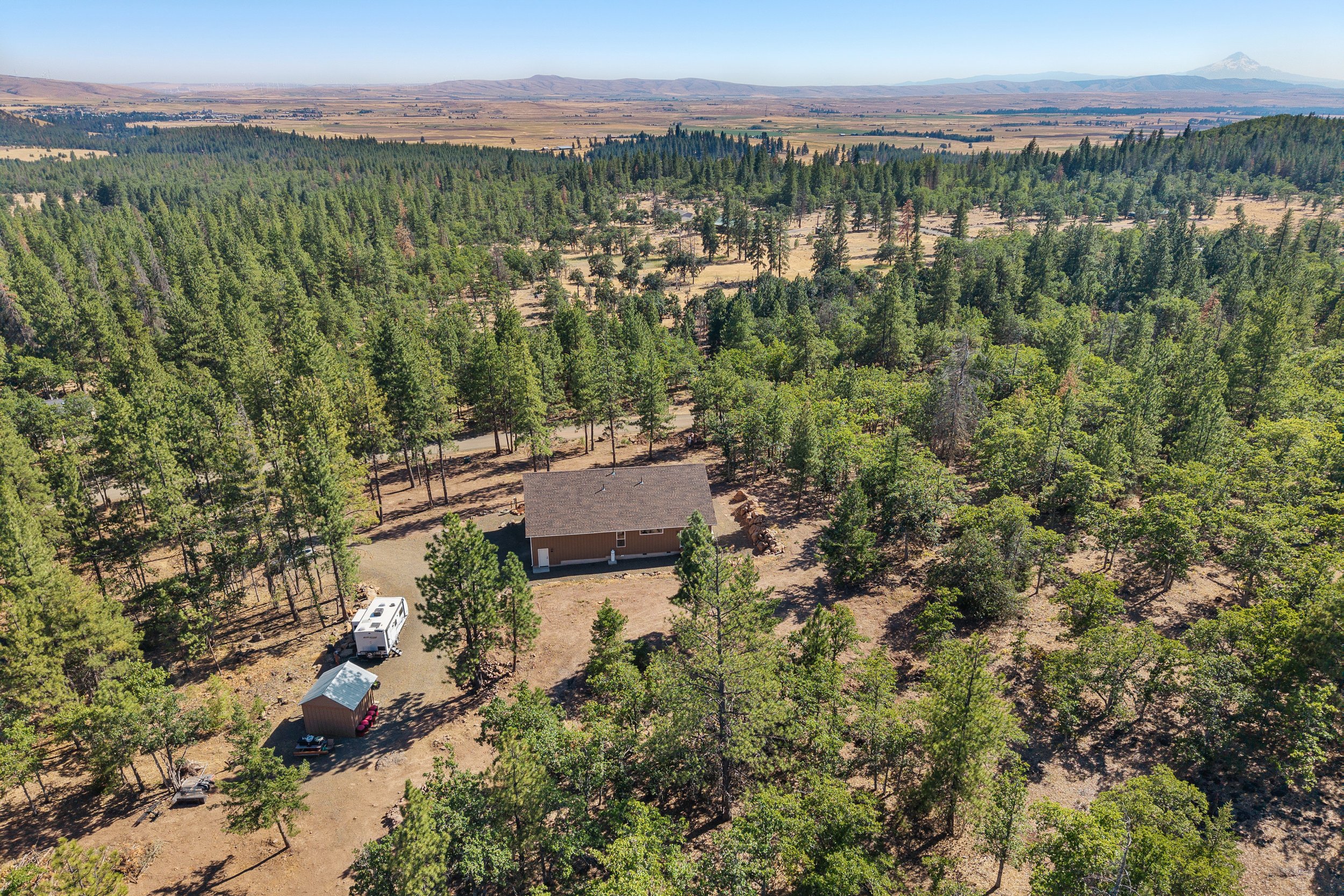 Aerial view of a house surrounded by trees in a rural landscape with mountains in the distance under a clear blue sky.