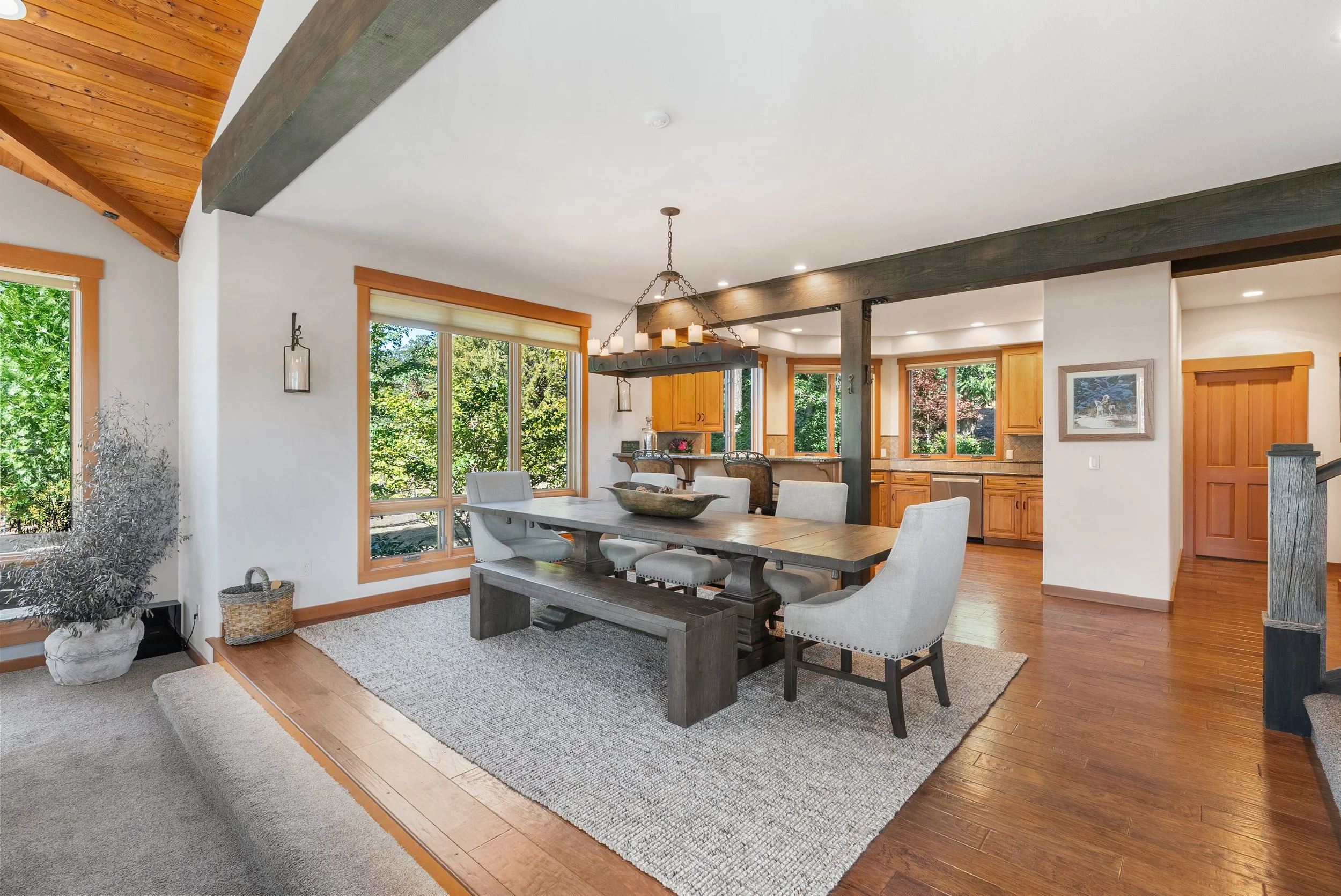 Open concept dining area with wooden table, upholstered chairs, large windows, and view of green trees outside.