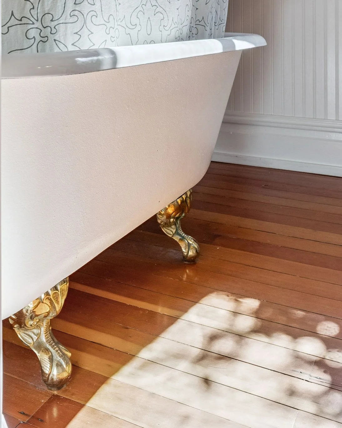 Close-up of a vintage clawfoot bathtub with ornate gold-colored feet on a wooden floor next to a wall with decorative paneling.