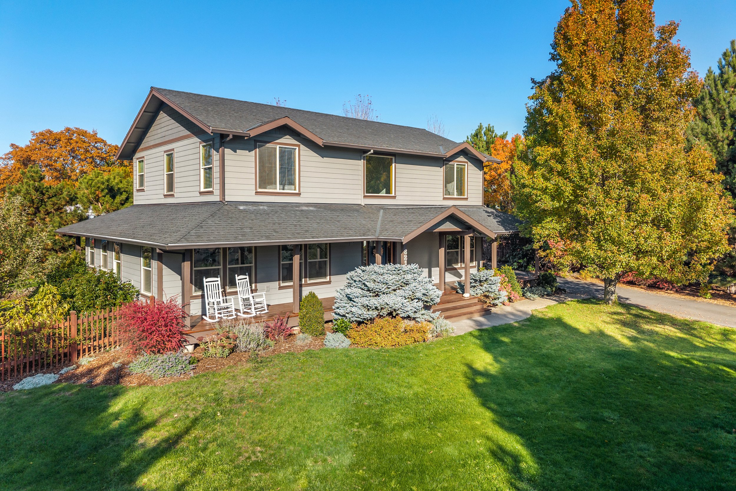 A two-story house with gray siding, brown trim, and a dark shingle roof, surrounded by green grass and colorful autumn trees.
