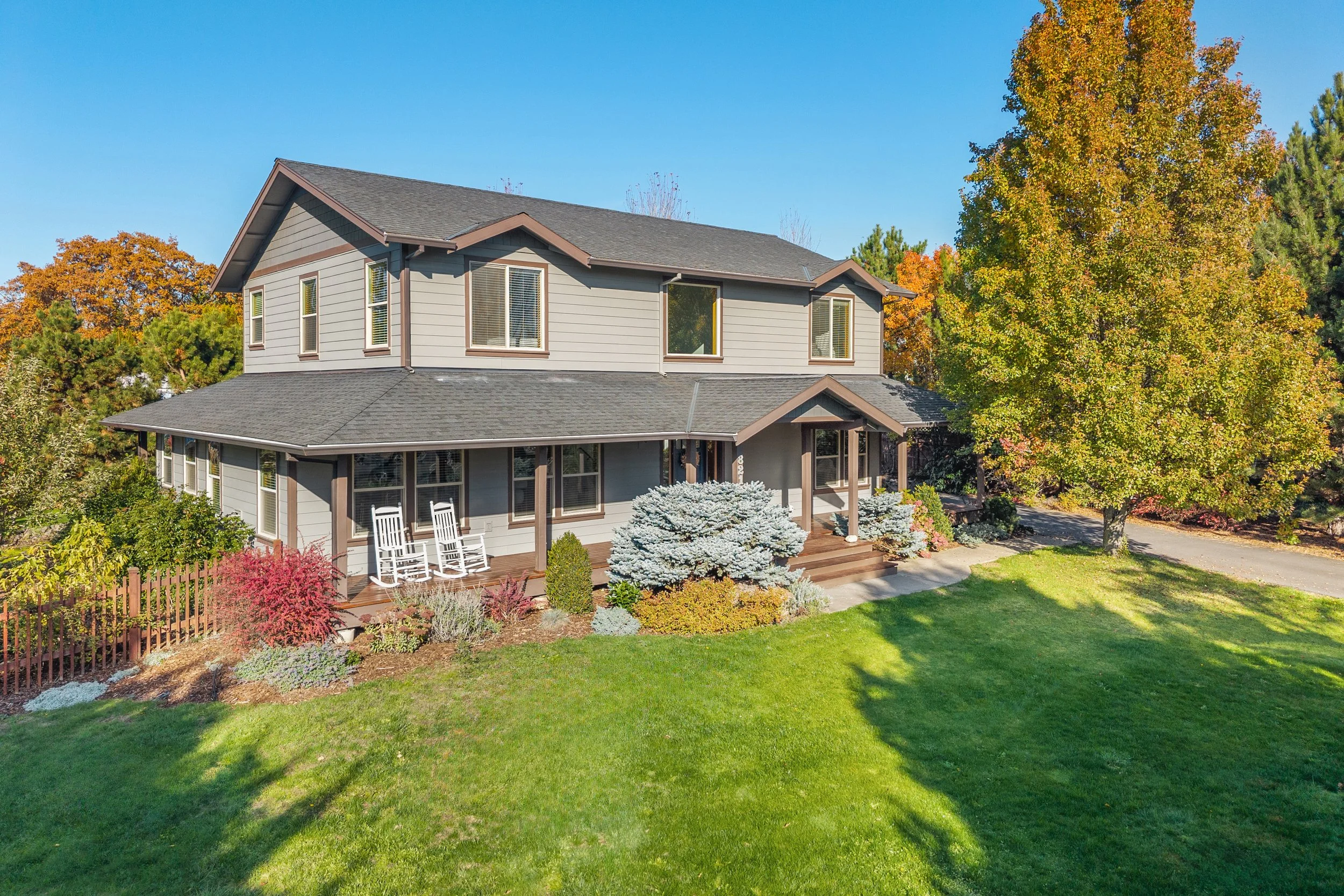 A two-story gray house with a covered front porch, white rocking chairs, surrounded by green lawn and colorful autumn trees, under a clear blue sky.