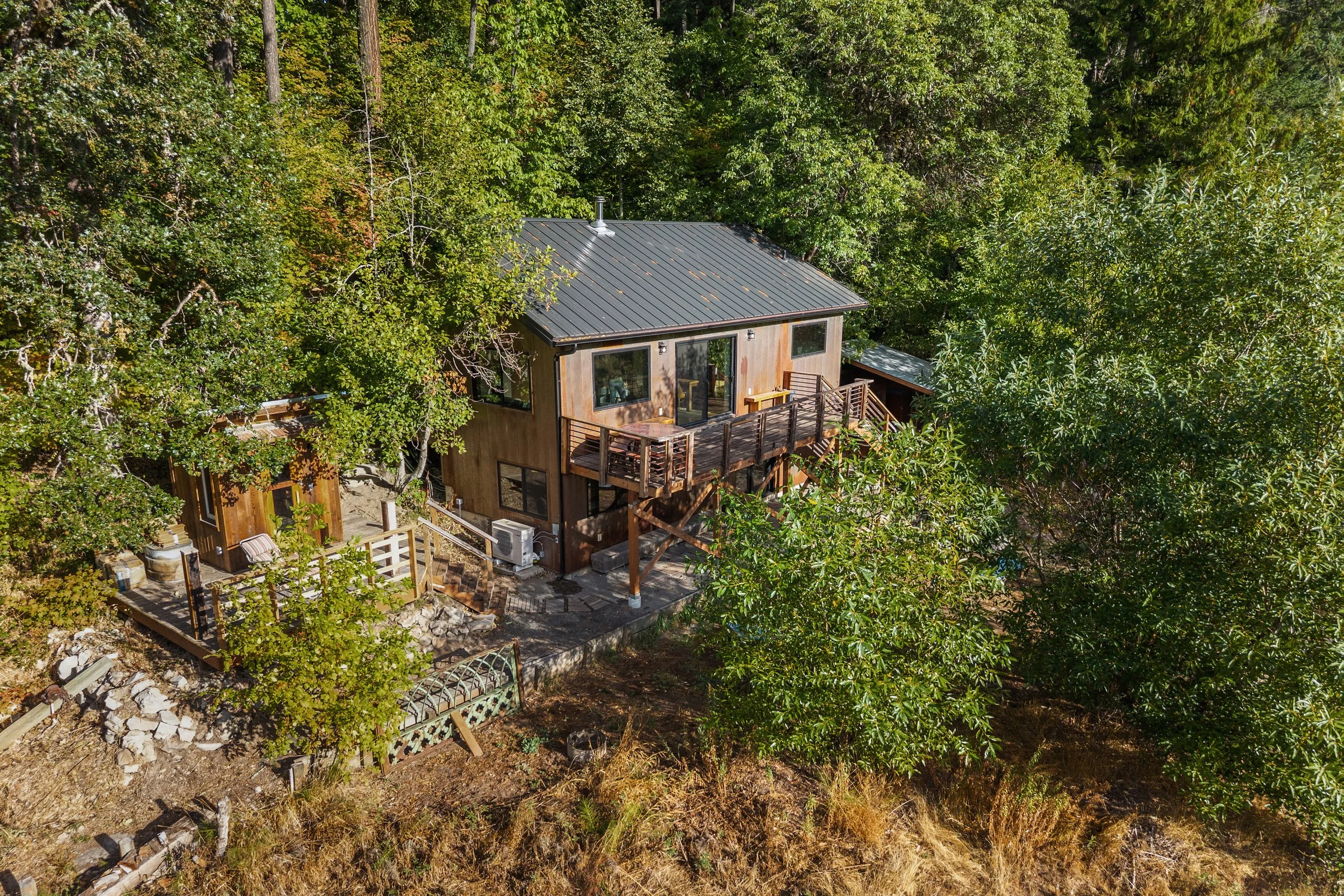 A two-story wooden house with a metal roof, surrounded by tall green trees, with a deck on the upper level and a patio area below.
