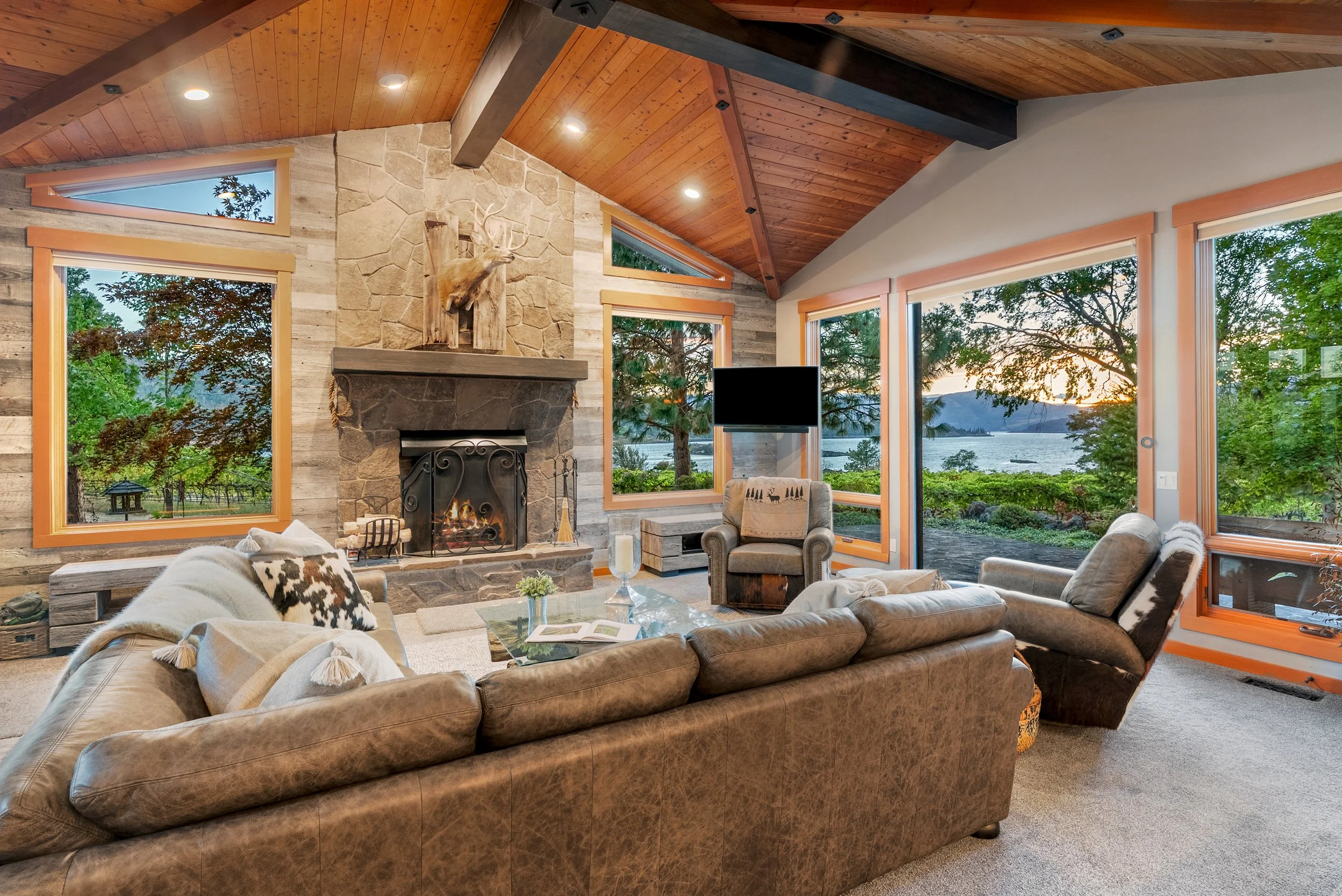 Living room with large windows showcasing a lakeside view, stone fireplace with mounted deer head, leather couch, armchairs, and a coffee table, during sunset.