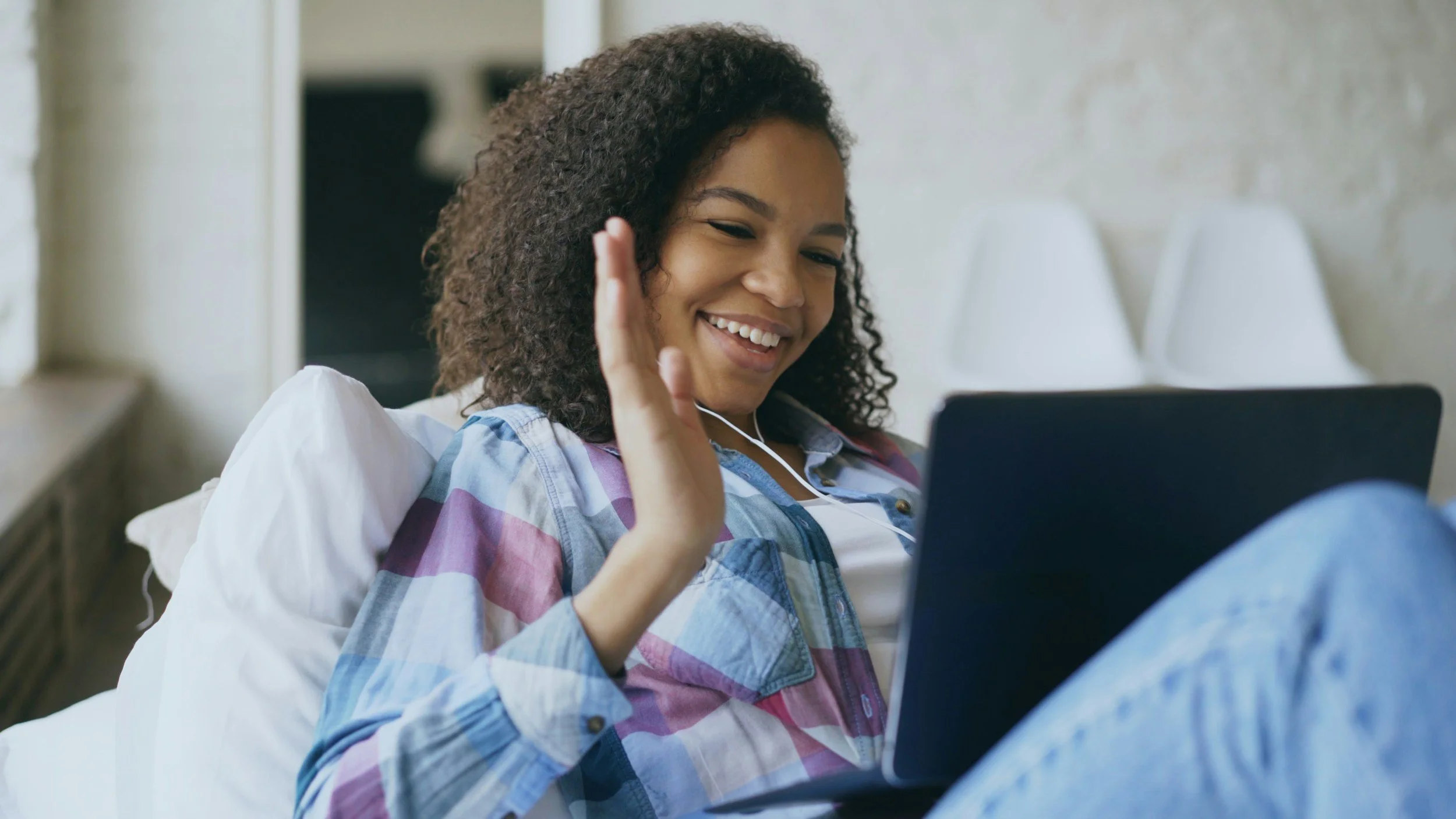 A teenage girl with curly hair smiling and waving while watching something on her laptop