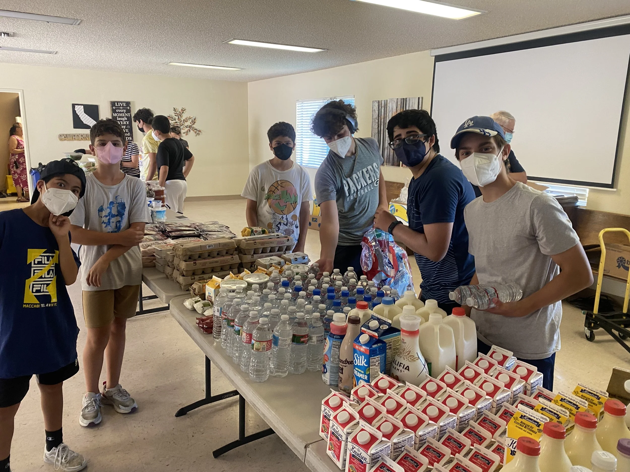 Group of teen boys wearing masks doing community service in a community center, with other people and tables in the background.