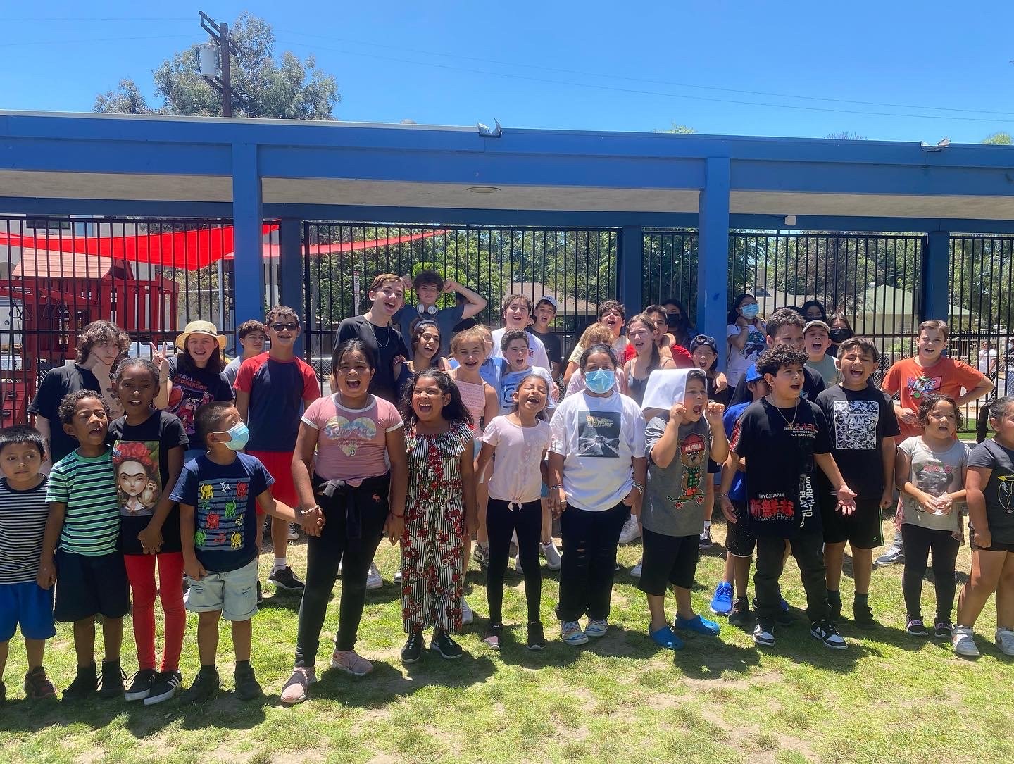 A group of children and a few adults gathered outside on a sunny day near a blue fence, with some children smiling, laughing, and talking. Some children are wearing masks, and there are colorful umbrellas and trees in the background.