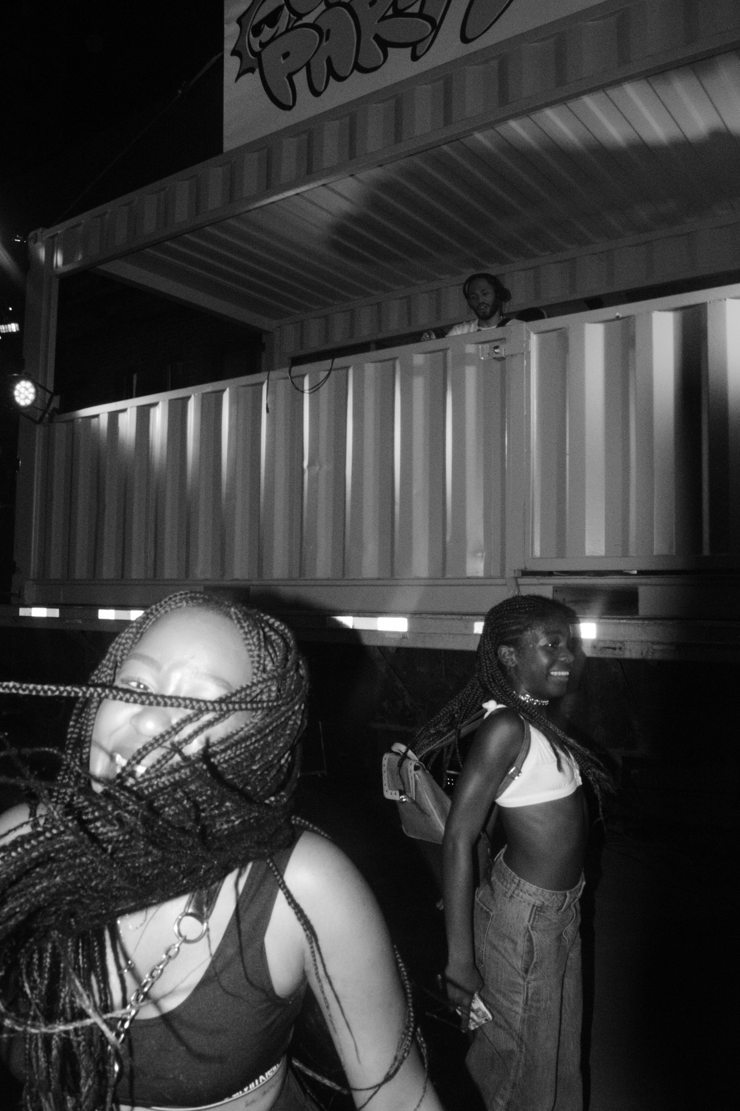 Two women with braided hair smiling in front of a large truck at night, with a DJ on a raised platform above them.