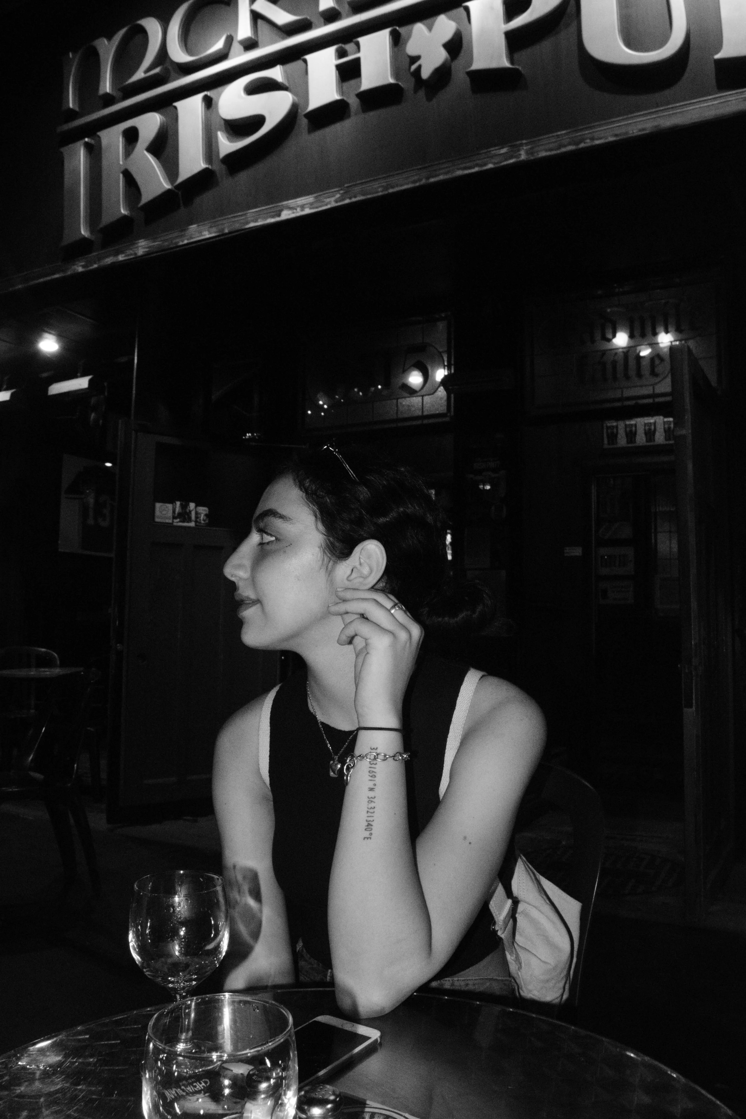 A young woman sitting at a table in a dimly lit restaurant, looking to her left, with glasses and a smartphone on the table, and a sign that says 'Tropicana Irish Pub' in the background.