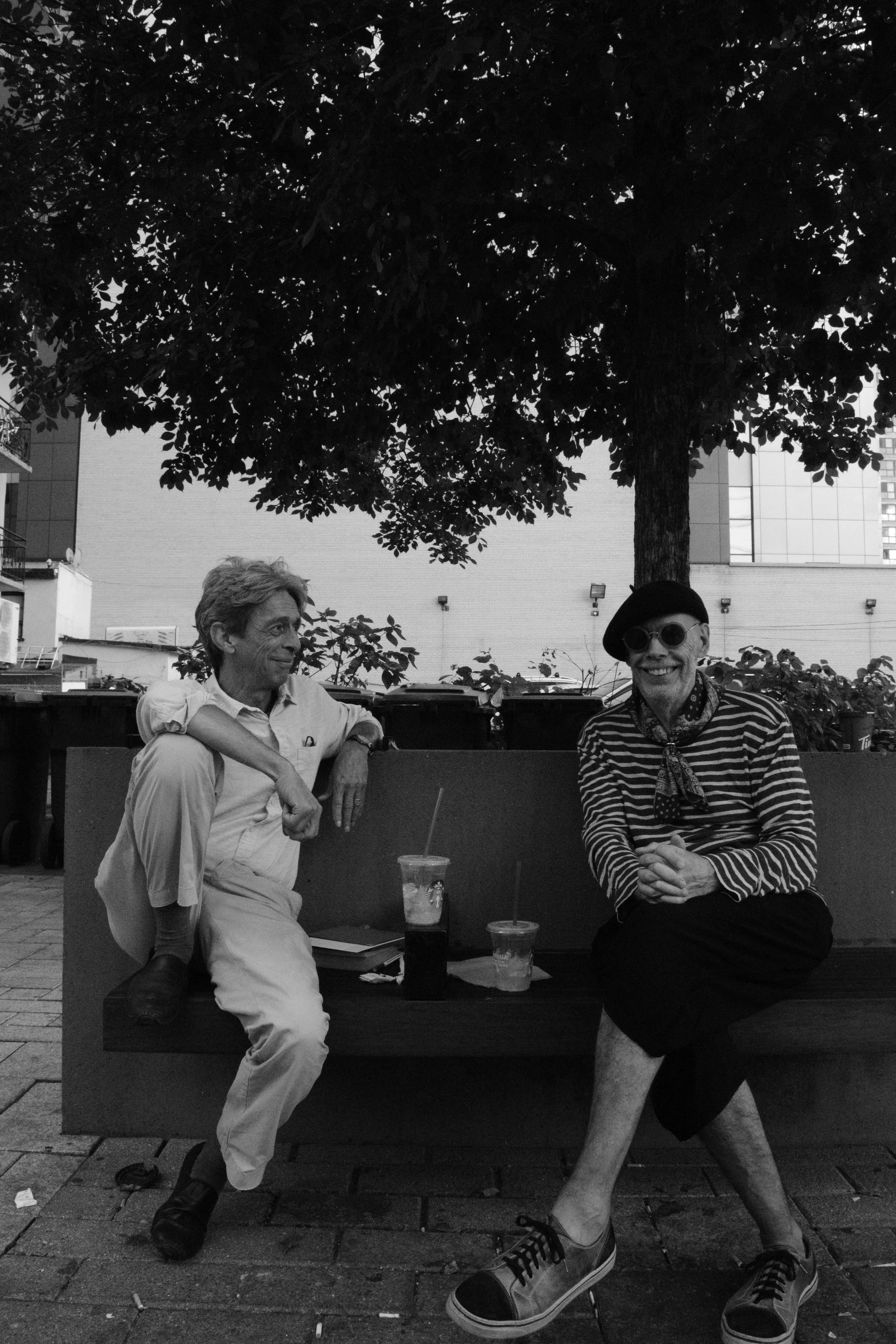 Two people sit on a bench under a large tree, smiling and talking, with drinks on the table, in an outdoor urban setting.