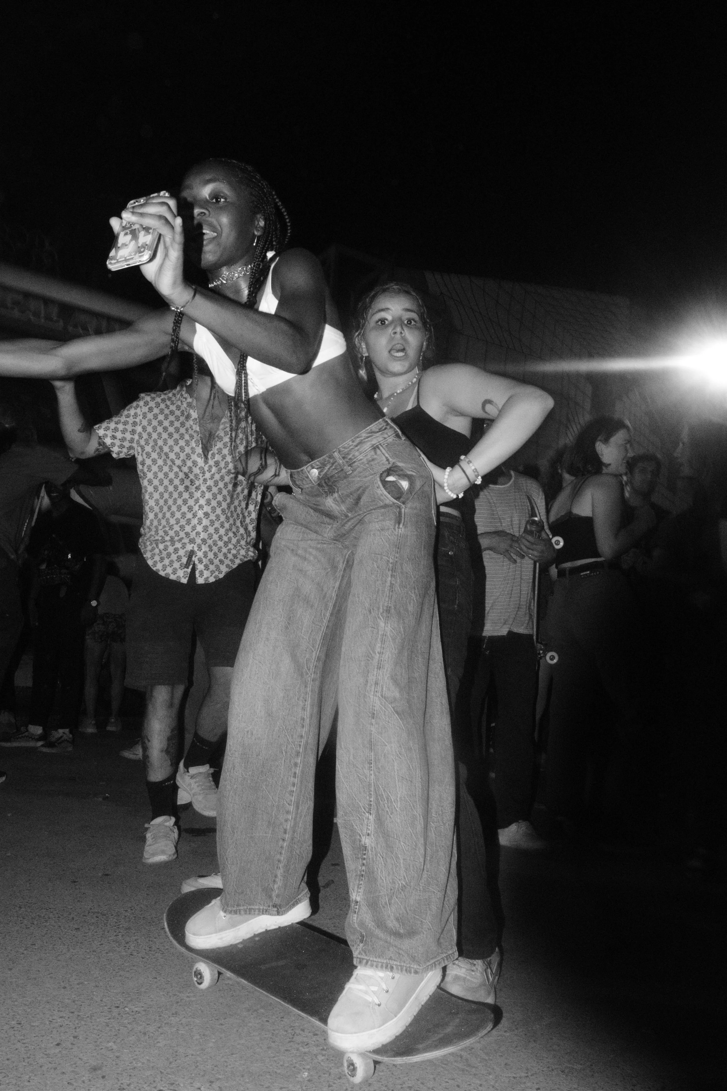 Two women skateboarding at night, one with braided hair taking a selfie, the other with short hair and tattoos, surrounded by people at a social event.