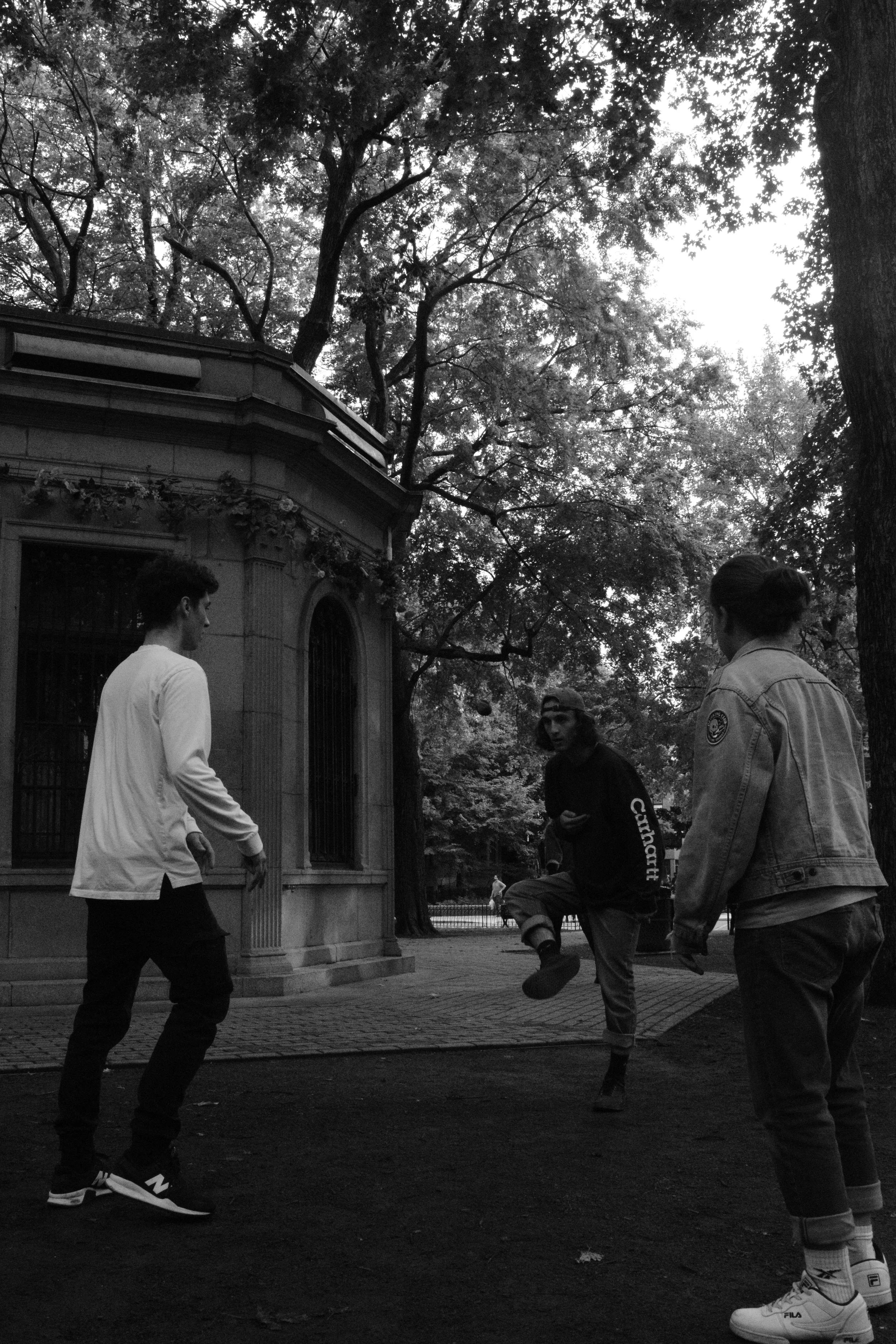 Three people playing with a ball in a park, with trees and a building in the background.