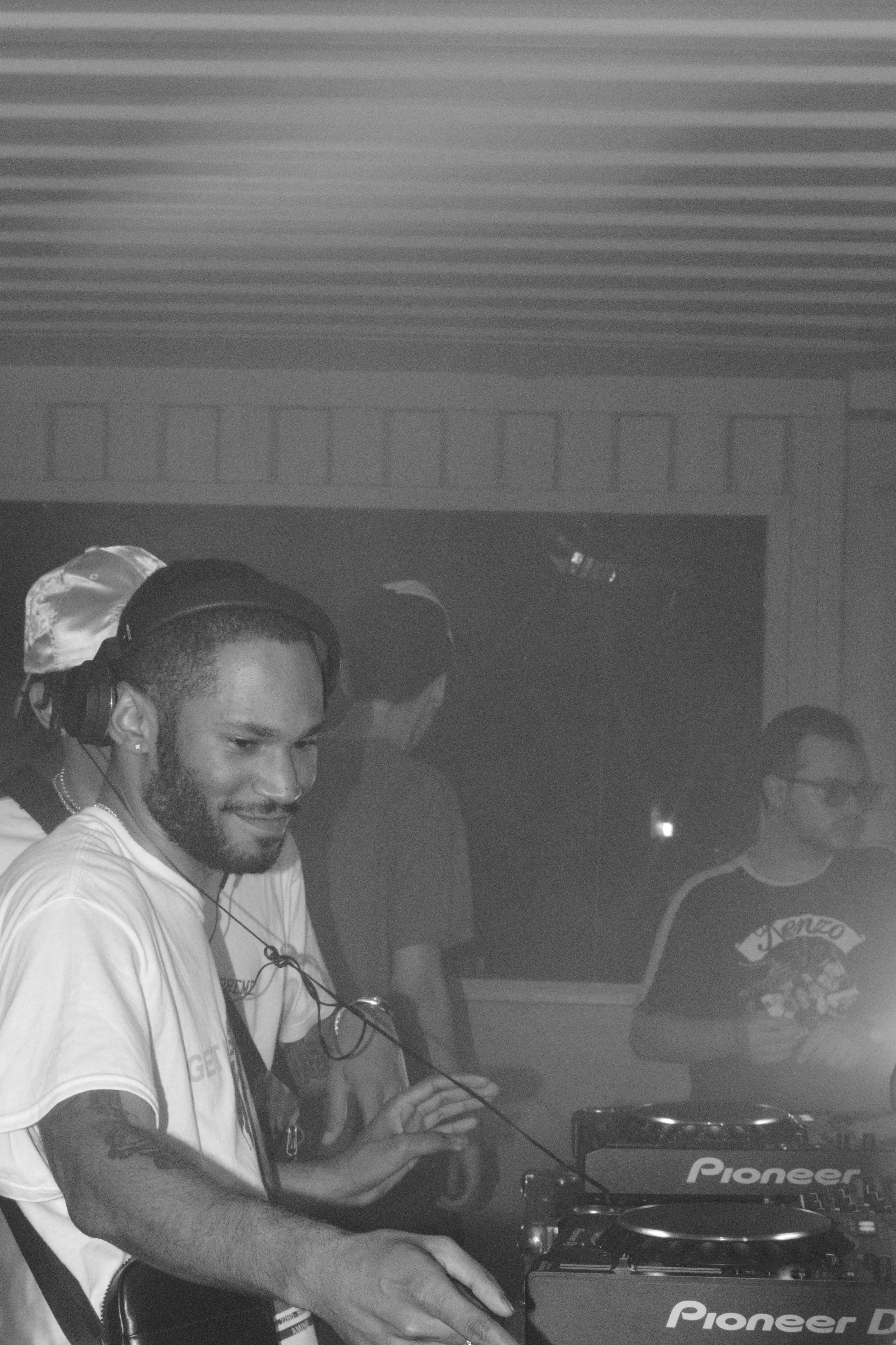 A DJ with headphones in a dark club or studio, mixing music on Pioneer DJ equipment, with people in the background.