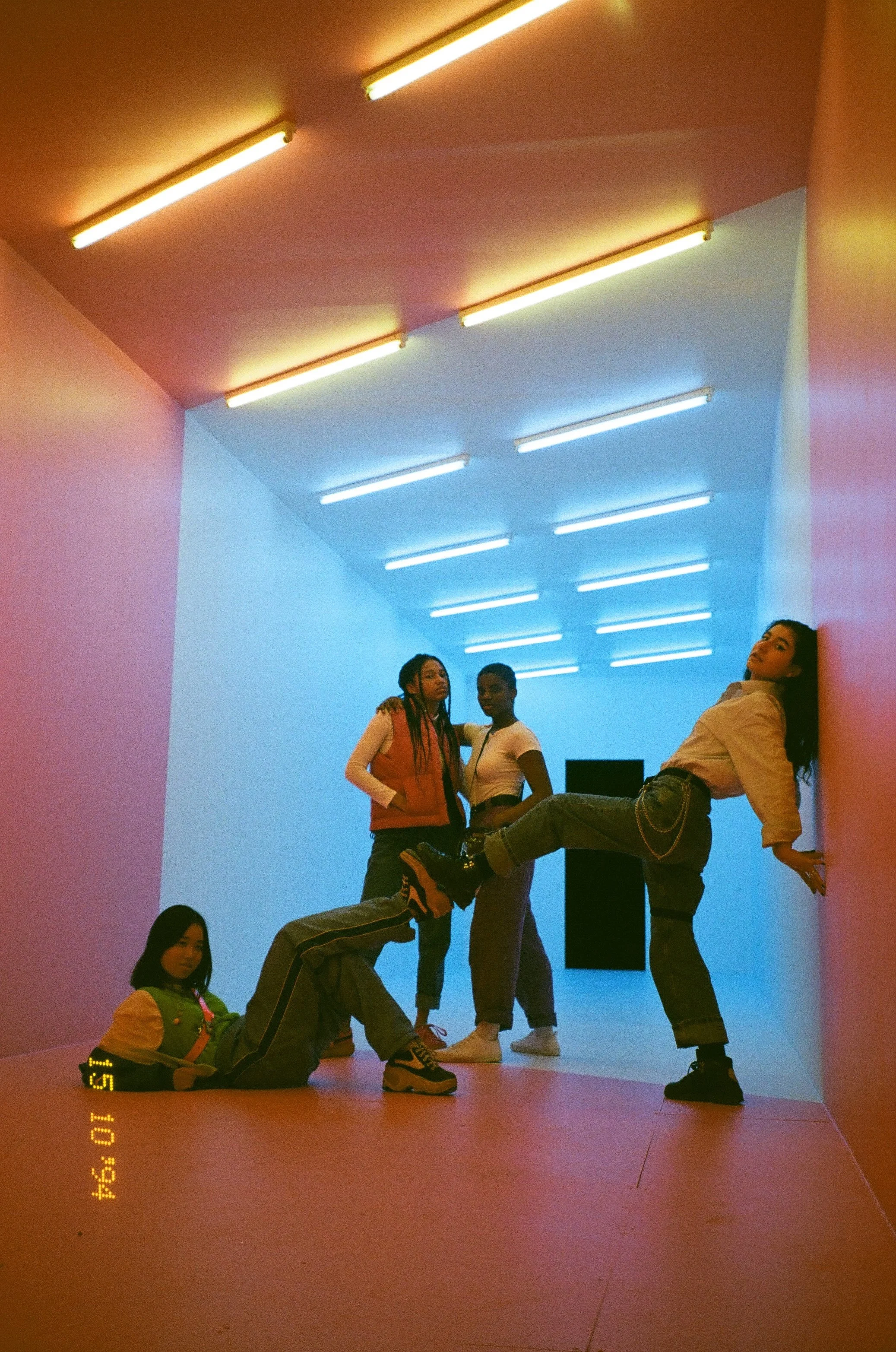 Four young women in a colorful, artistic room with many fluorescent lights on the ceiling. One woman is sitting on the floor, another is leaning against a pink wall with her leg raised, and two are standing in the background.